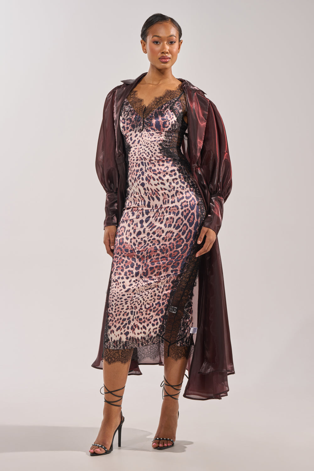 A woman stands confidently against a plain light background, wearing the PRETTY LIL THING ORGANZA TRENCH—an elegant sheer brown coat styled over a leopard print lace slip dress and black strappy heels. 5
