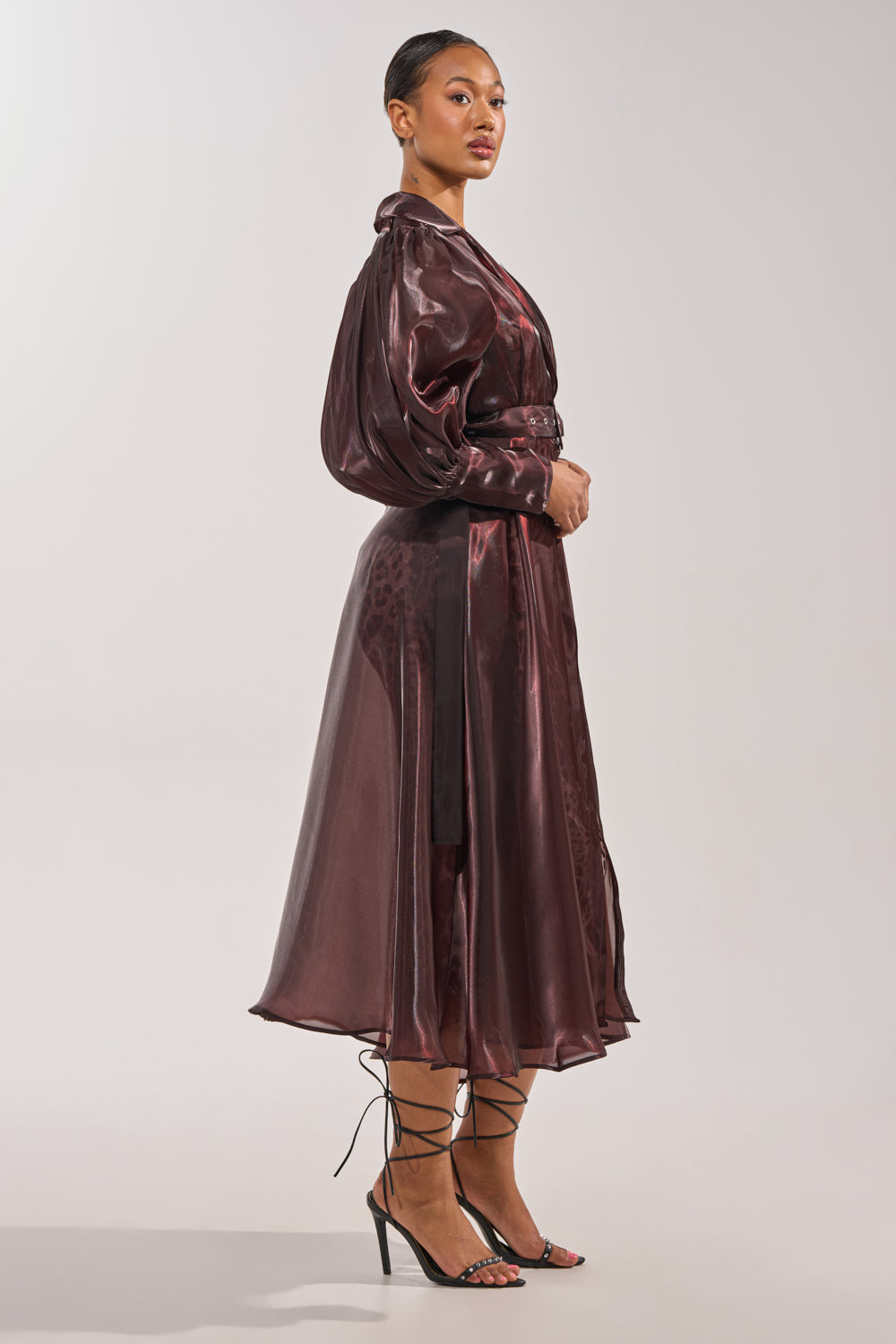 A woman in profile wears a shiny burgundy belted midi dress, strappy black heels, and her hair pulled back. She completes the look with the PRETTY LIL THING ORGANZA TRENCH over a light, plain background. 6