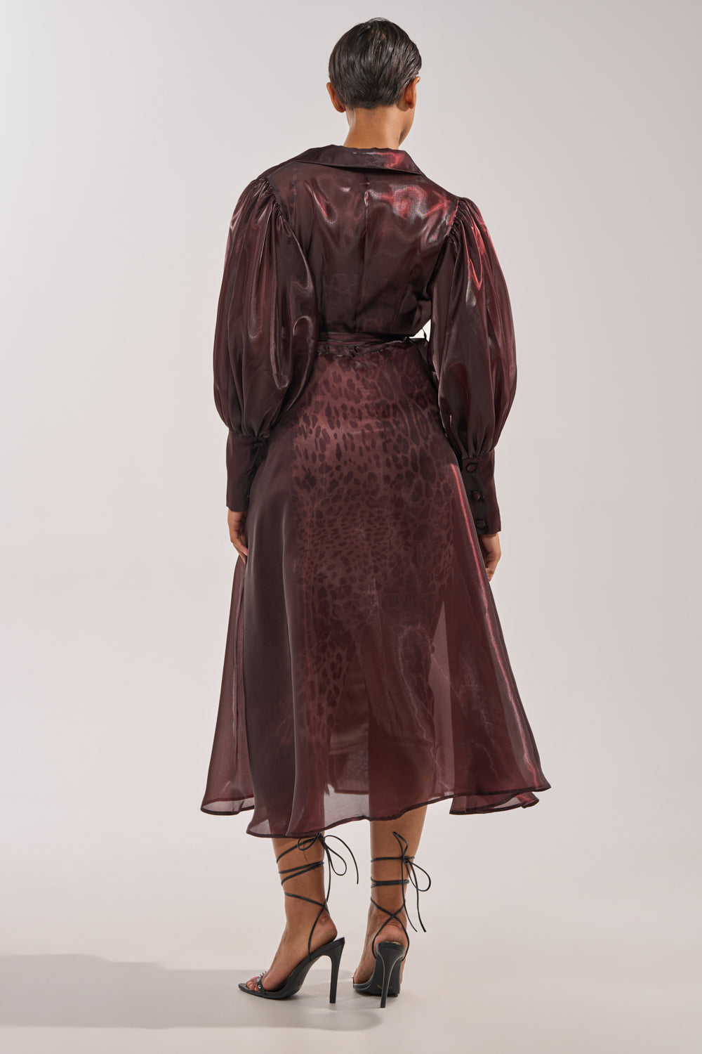 A person with short hair faces away, layering the PRETTY LIL THING ORGANZA TRENCH—sheer and brown—over a burgundy, puffed-sleeve, midi animal-print dress and finishing the look with black strappy high-heeled sandals. 7