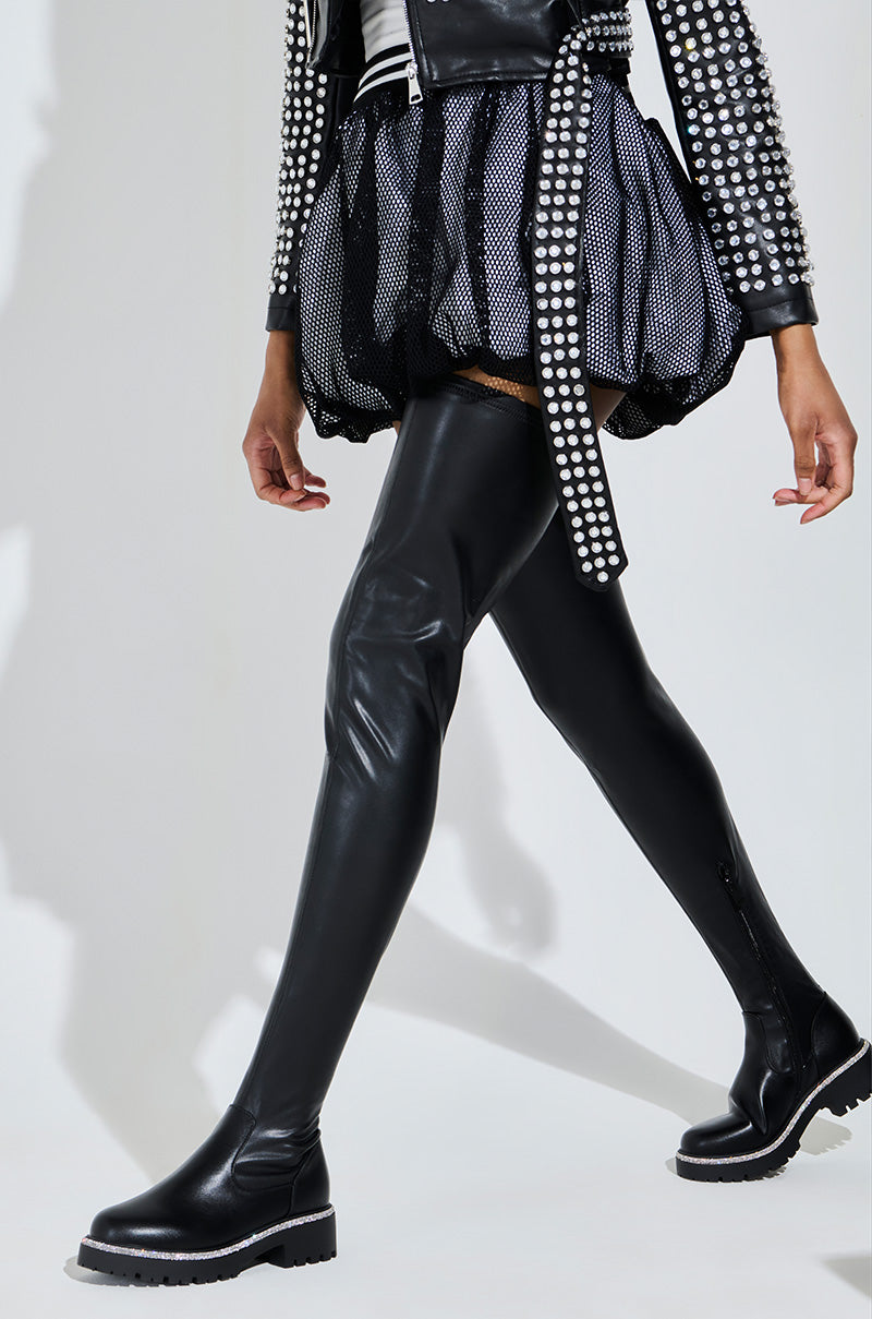 A person strides confidently in AZALEA WANG NEWRULES Rhinestone Stretch PU Flat Boots, paired with a black leather jacket adorned with silver studs and a black and white checkered skirt, standing out against a light background with their fashion-forward style.