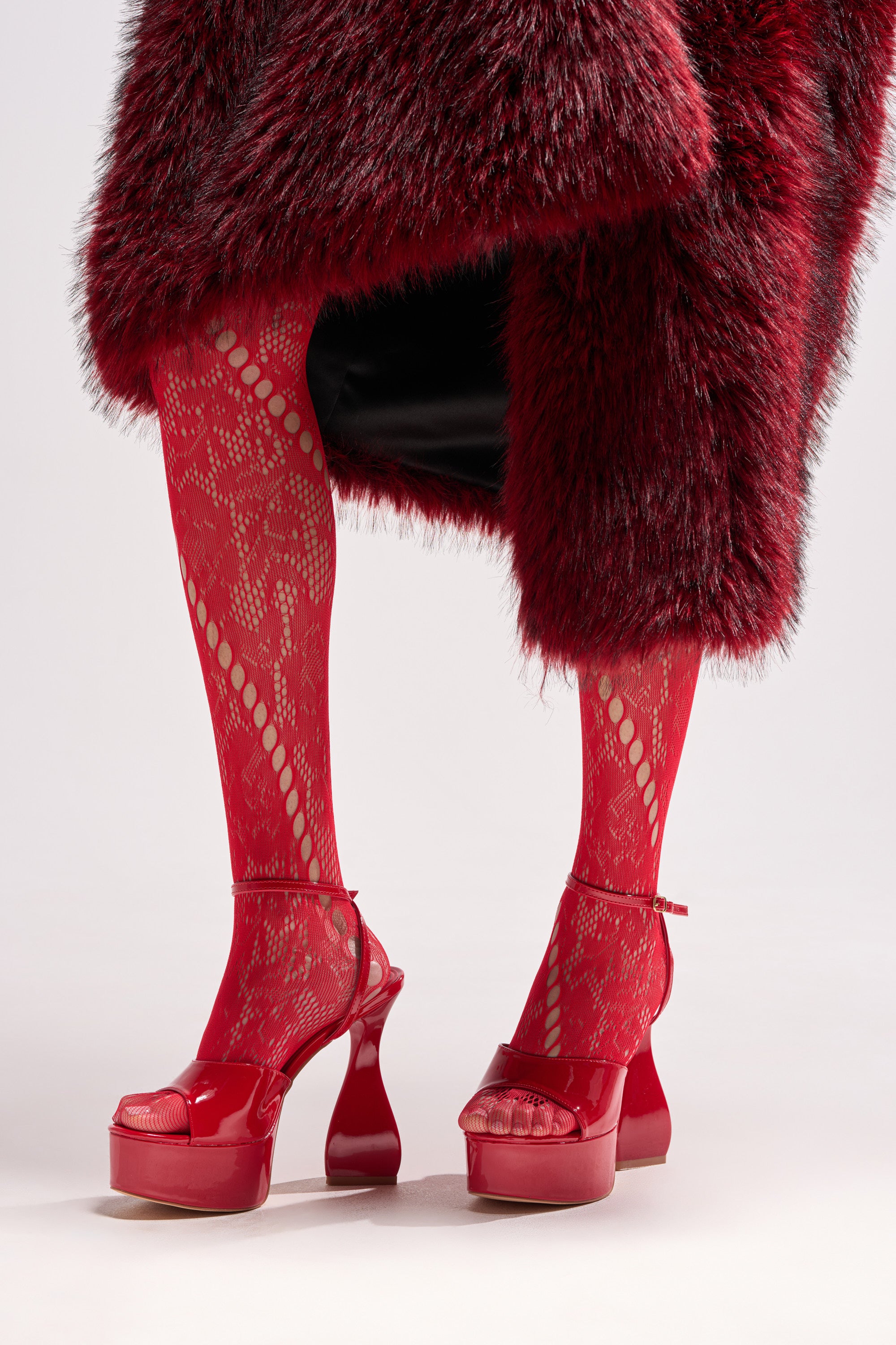 A person wearing patterned red lace tights, a knee-length dark red faux fur coat, and AZALEA WANG NEWTON RED CHUNKY PLATFORM SANDAL stands on a white background.