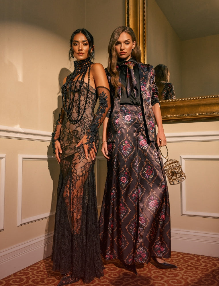Two models pose in elegant evening gowns in an indoor setting with a large gold mirror. The model on the left wears a sheer black lace halter-neck gown, while the model on the right wears a matching black and brown patterned long-sleeve dress with a high neck and bow tie collar, holding a small metallic handbag.