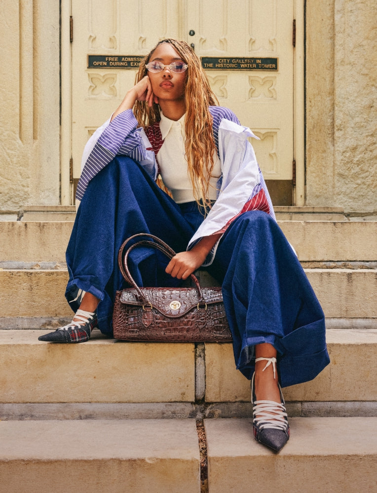 A woman sits on stone steps in front of a cream-colored building door, wearing a white and blue striped jacket, wide-leg navy pants, and lace-up pointed shoes, while holding a brown crocodile-embossed handbag.