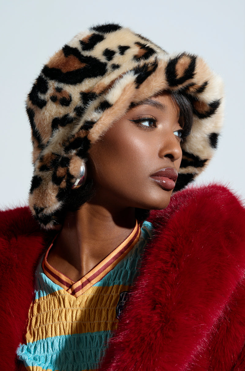 The individual wearing the SISTA SISTA CHEETAH BUCKET HAT and a red faux fur coat gazes to the side. They have large silver hoop earrings and sport a colorful top underneath, set against a soft, neutral background.