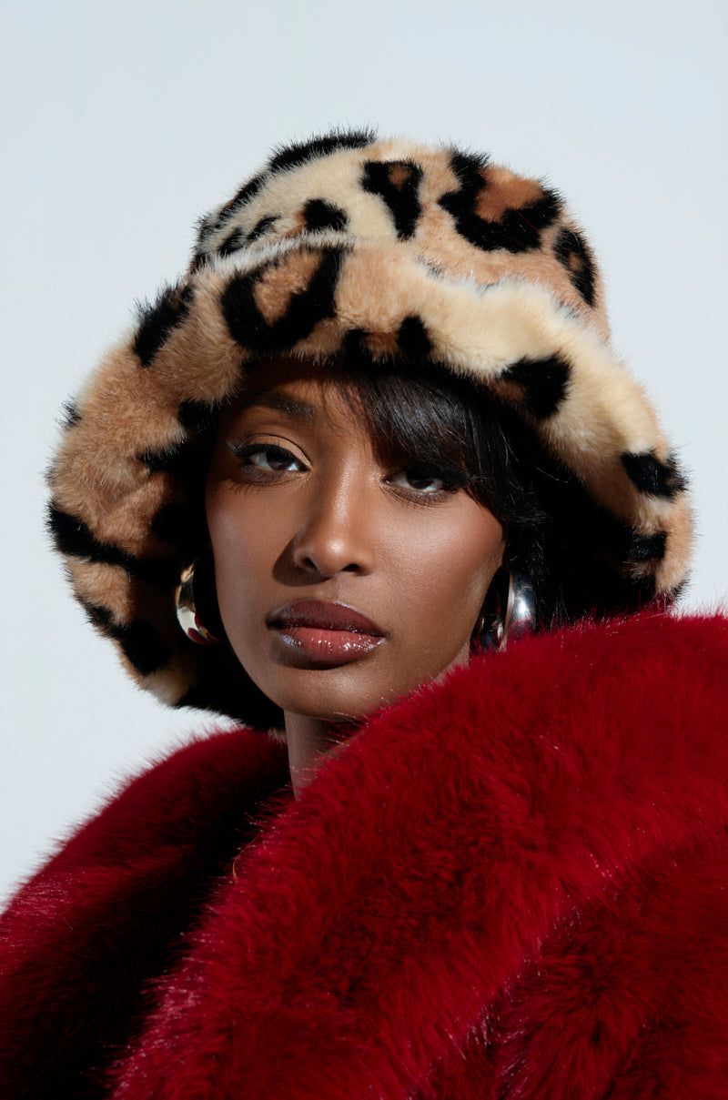 A person wearing the SISTA SISTA CHEETAH BUCKET HAT and a vibrant red faux fur coat stands with large hoop earrings, gazing directly at the camera against a plain background.