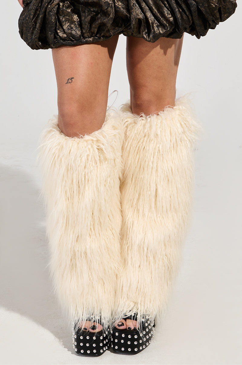 A person wearing the TUNDRA FAUX FUR THIGH WARMERS in a fluffy, cream color, paired with black platform sandals adorned with silver studs. The outfit is completed with a textured skirt that has a metallic appearance, and on one thigh, there's a tattoo displaying the number "15." The thigh warmers suggest an inner sherpa lining for extra comfort.