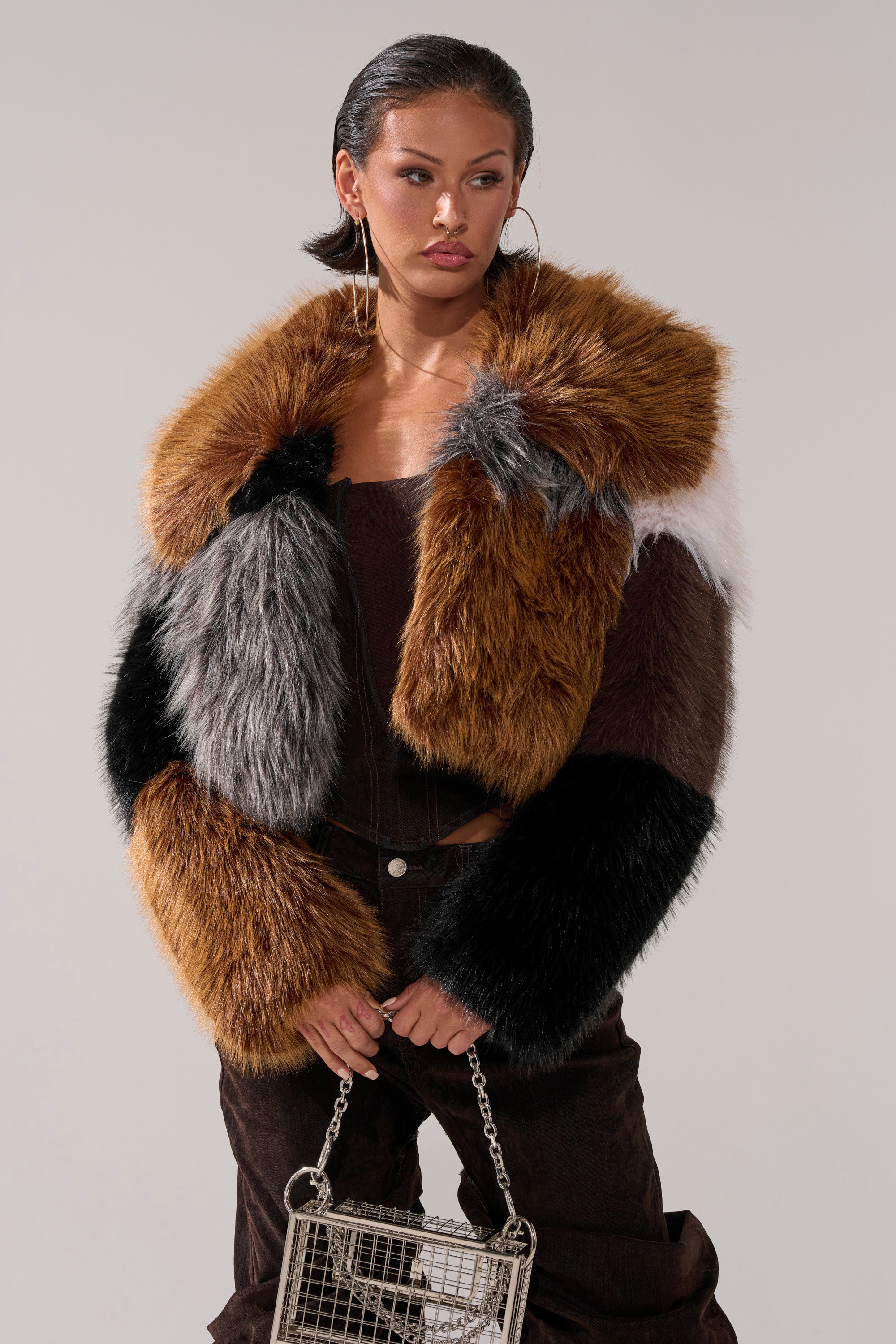 A woman stands against a plain background in the HAZEL PATCHWORK FAUX FUR COAT, featuring shades of brown, black, gray, and white. She completes the look with a small silver chain handbag, slicked-back hair, and hoop earrings.