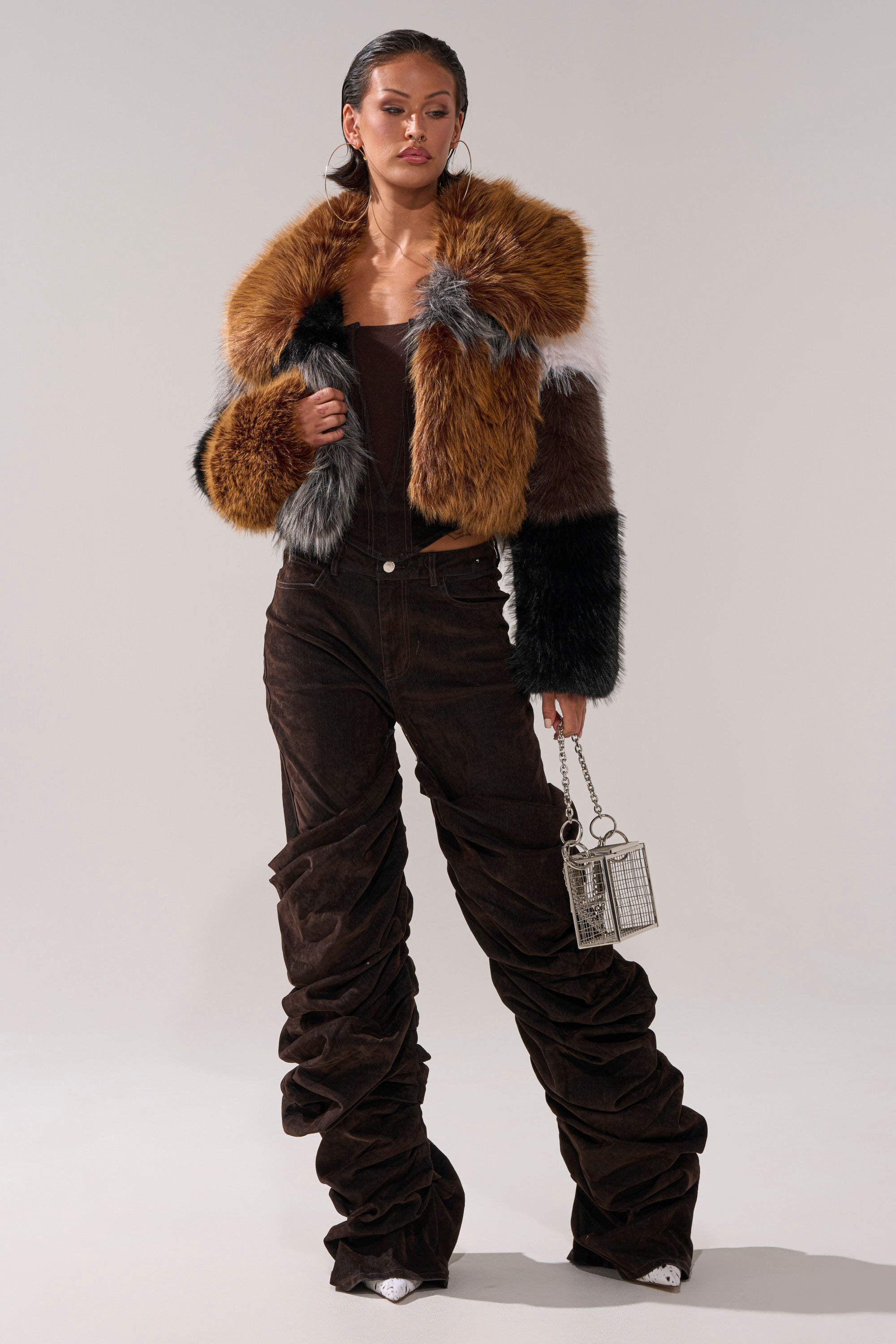 A woman poses in the HAZEL PATCHWORK FAUX FUR COAT, featuring brown, orange, and gray tones. She pairs it with high-waisted brown pants, white heels, a small silver handbag, and hoop earrings.