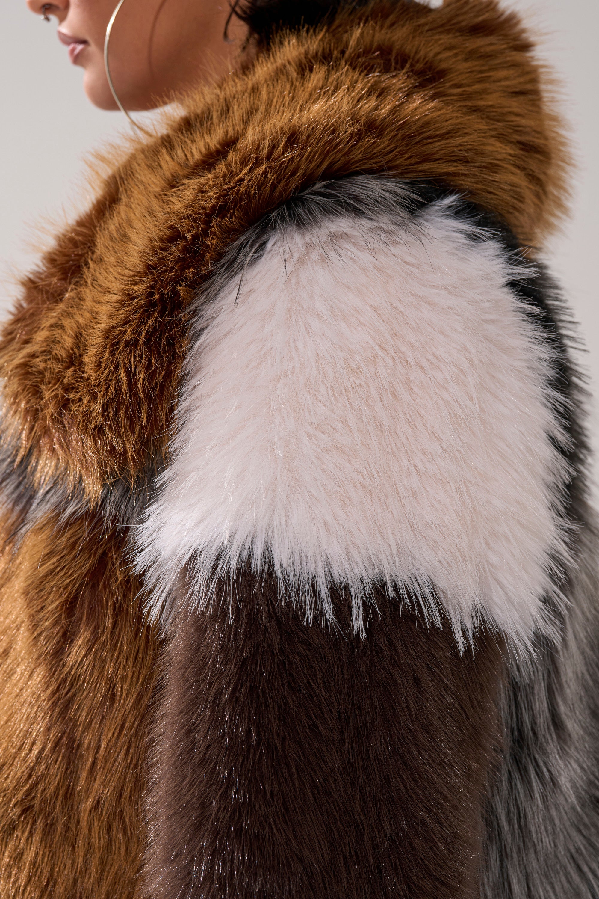 Close-up of a person wearing the HAZEL PATCHWORK FAUX FUR COAT, featuring soft, fluffy brown, white, and gray sections. The person's face is partially visible on the left.