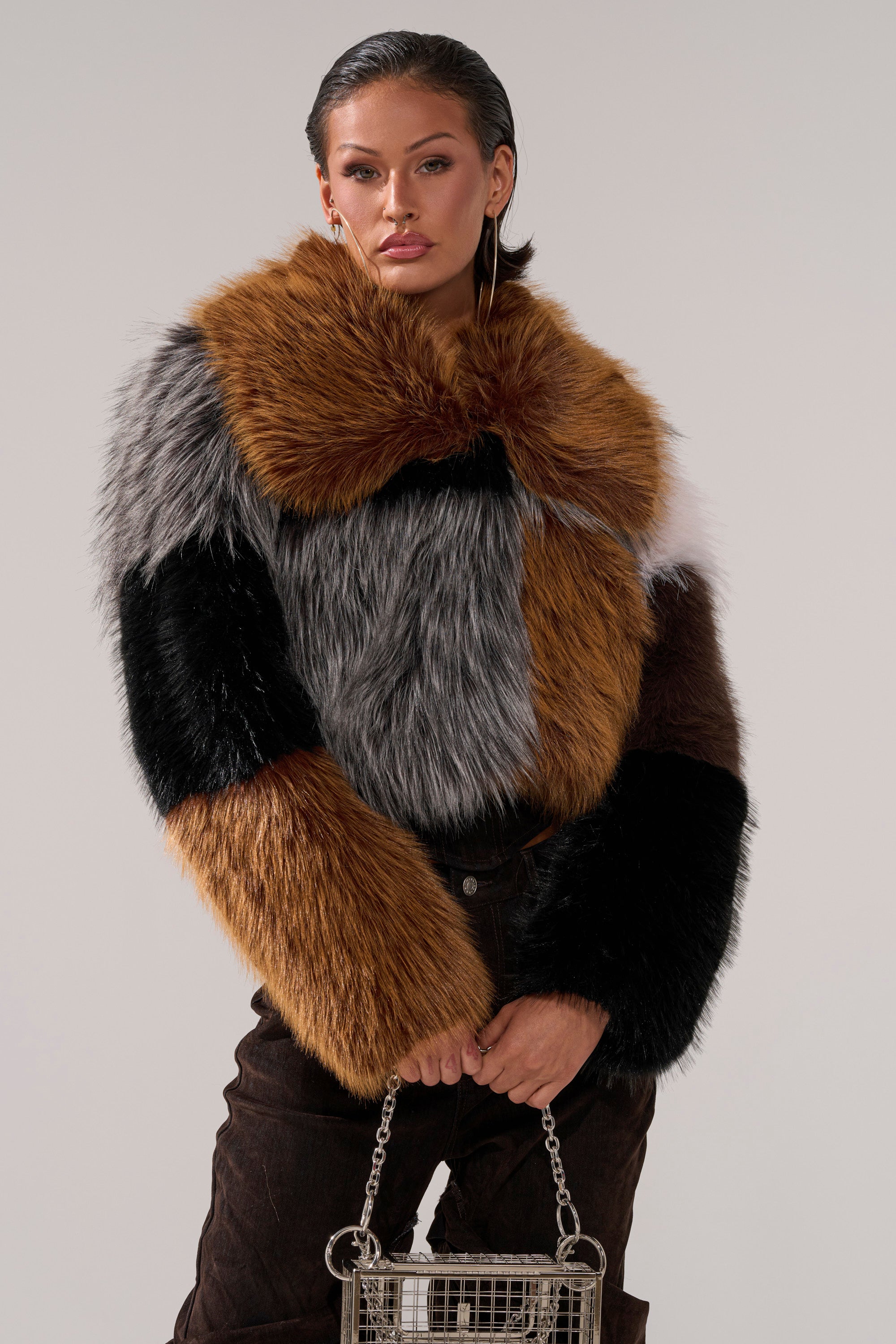 A woman models the HAZEL PATCHWORK FAUX FUR COAT with brown, black, and gray patches, paired with a silver metal handbag. She has slicked-back hair, hoop earrings, and poses confidently against a plain background.