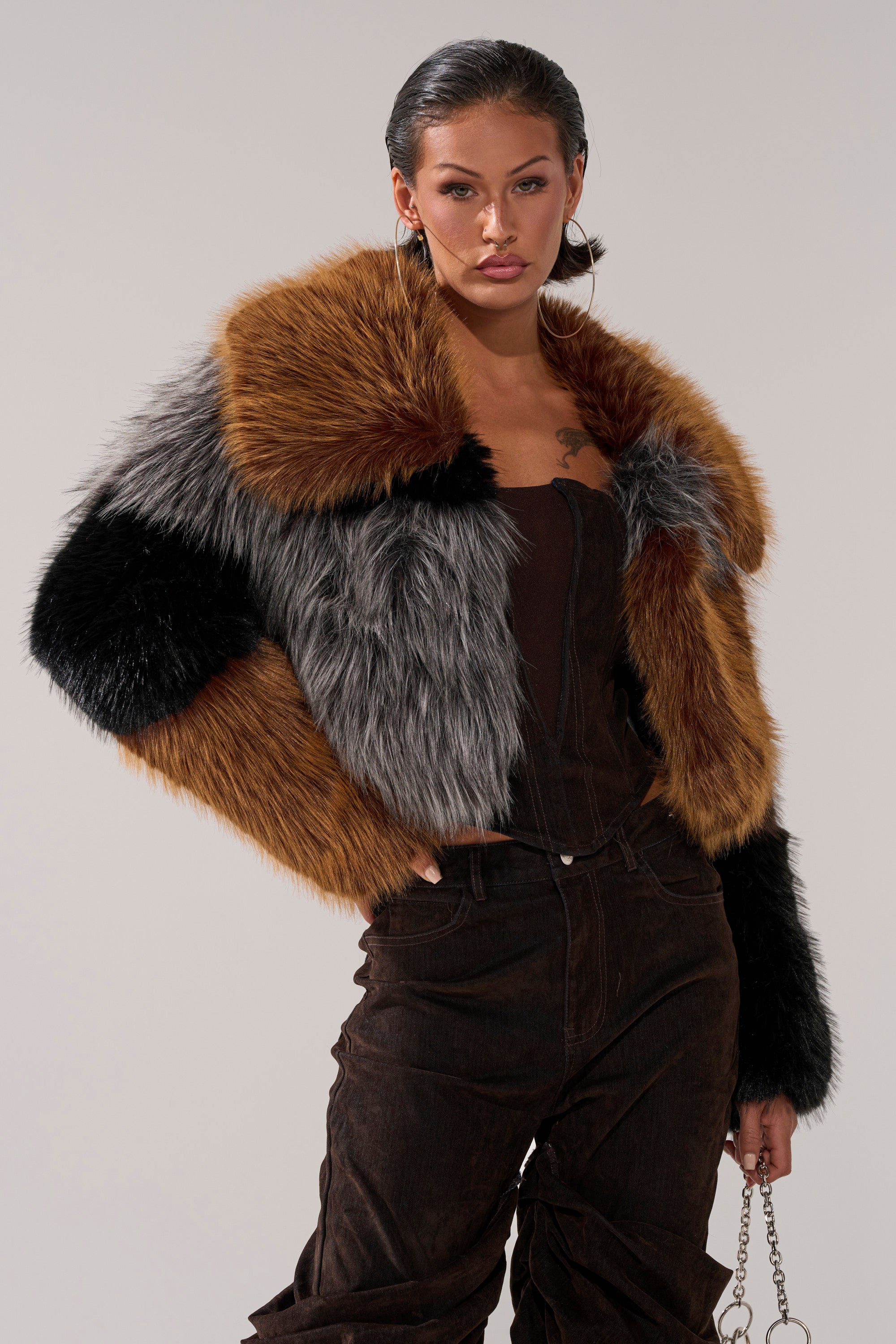 A woman with slicked-back hair models the HAZEL PATCHWORK FAUX FUR COAT in brown, grey, and black over a dark top and brown pants, posing confidently with one hand on her hip against a plain background.