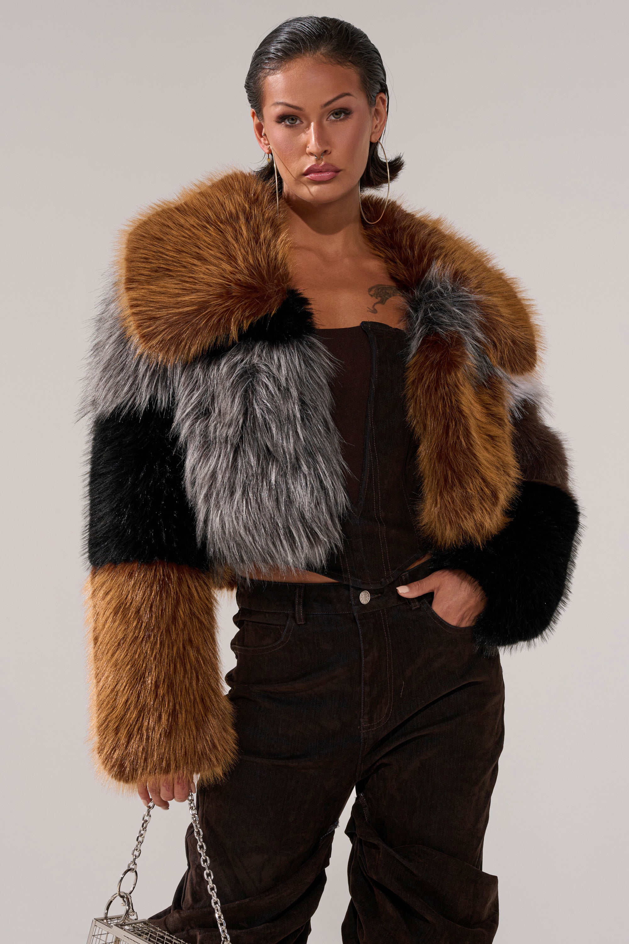 A woman with slicked-back hair wears large hoop earrings, the HAZEL PATCHWORK FAUX FUR COAT in brown, black, and gray over a dark top and pants, and holds a small silver purse.