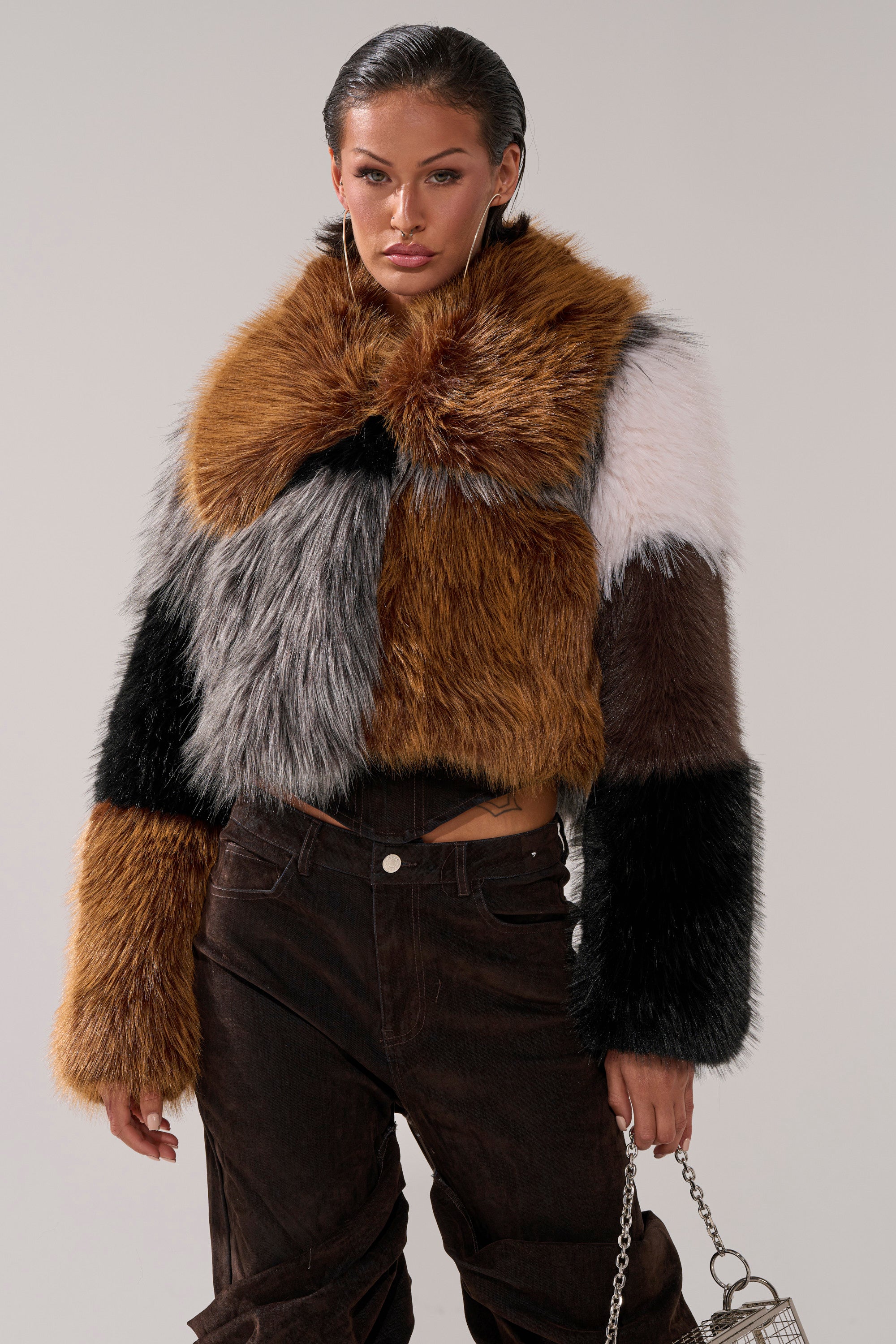 A woman stands against a plain background wearing the HAZEL PATCHWORK FAUX FUR COAT in brown, black, white, and gray, paired with dark high-waisted pants and a small silver handbag.
