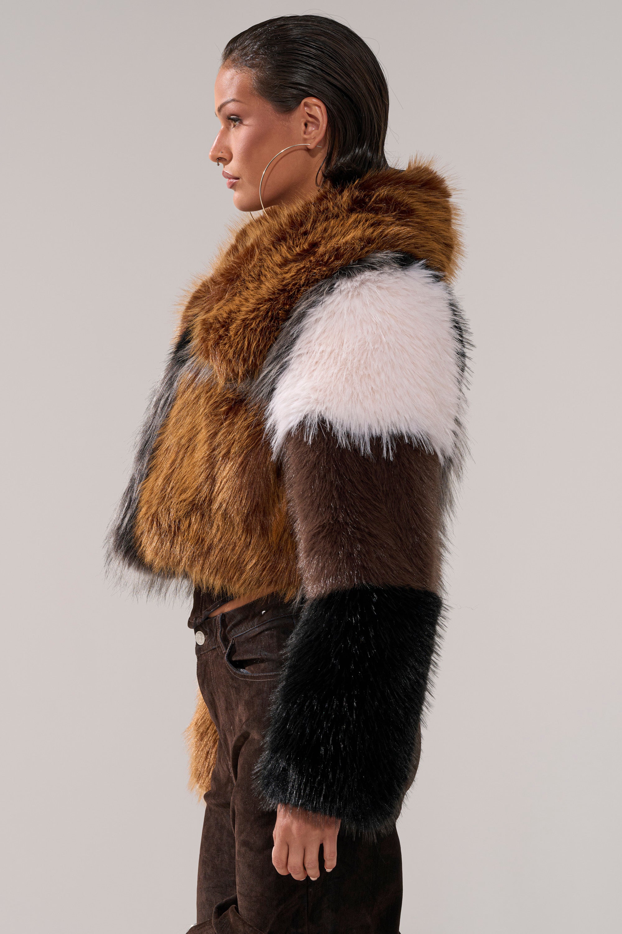 A woman with slicked-back hair models the HAZEL PATCHWORK FAUX FUR COAT in brown, black, white, and rust, paired with dark pants. She stands sideways against a plain light background.