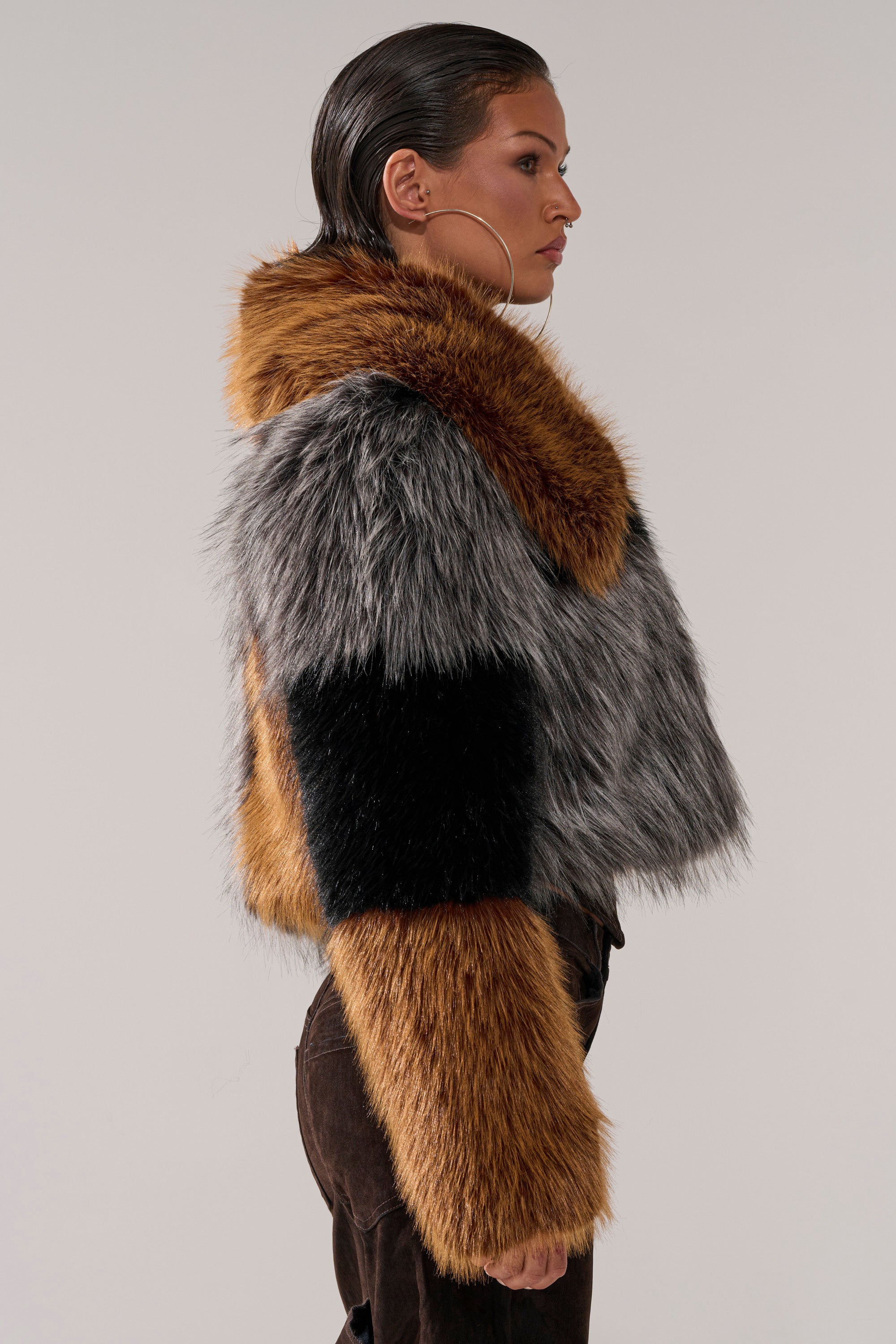 A woman in profile wears the HAZEL PATCHWORK FAUX FUR COAT, featuring brown, black, and gray sections. She pairs it with dark pants, large hoop earrings, and slicked-back hair against a plain light background.