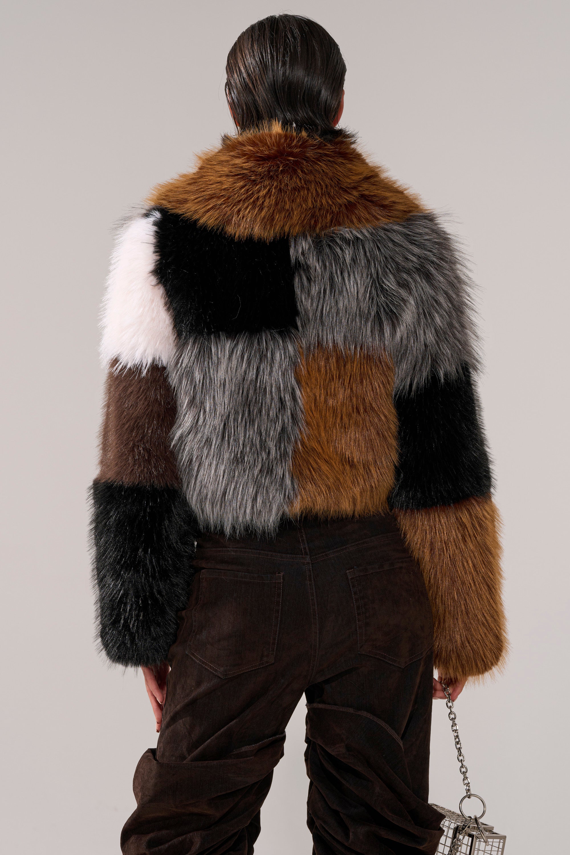 A person stands with their back to the camera, wearing the HAZEL PATCHWORK FAUX FUR COAT in brown, black, white, and gray shades, paired with dark pants and holding a shiny handbag.