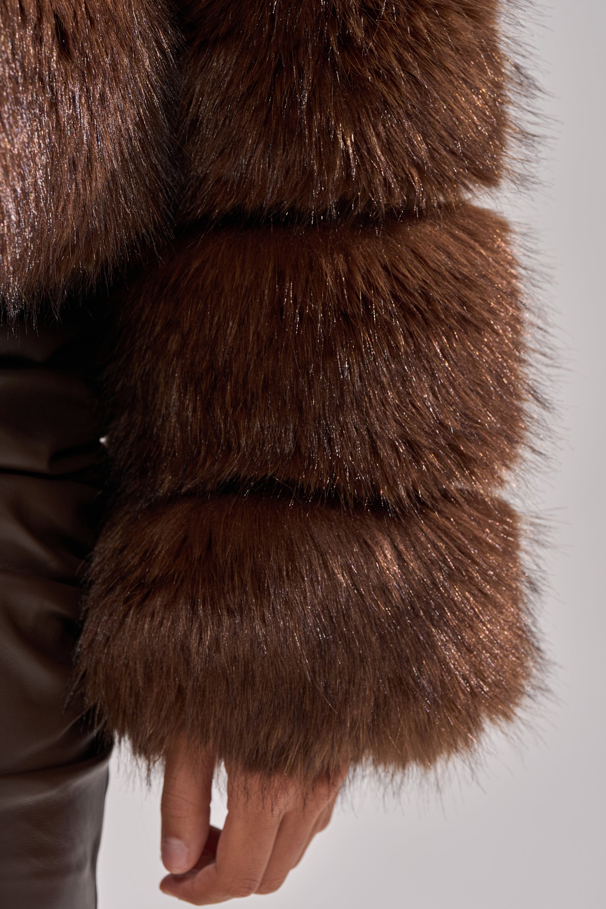 A close-up of a person’s arm wearing the GOOD KARMA FAUX FUR jacket, featuring thick brown sections and an oversized collar, paired with dark leather pants. The relaxed hand is partially visible at the bottom.