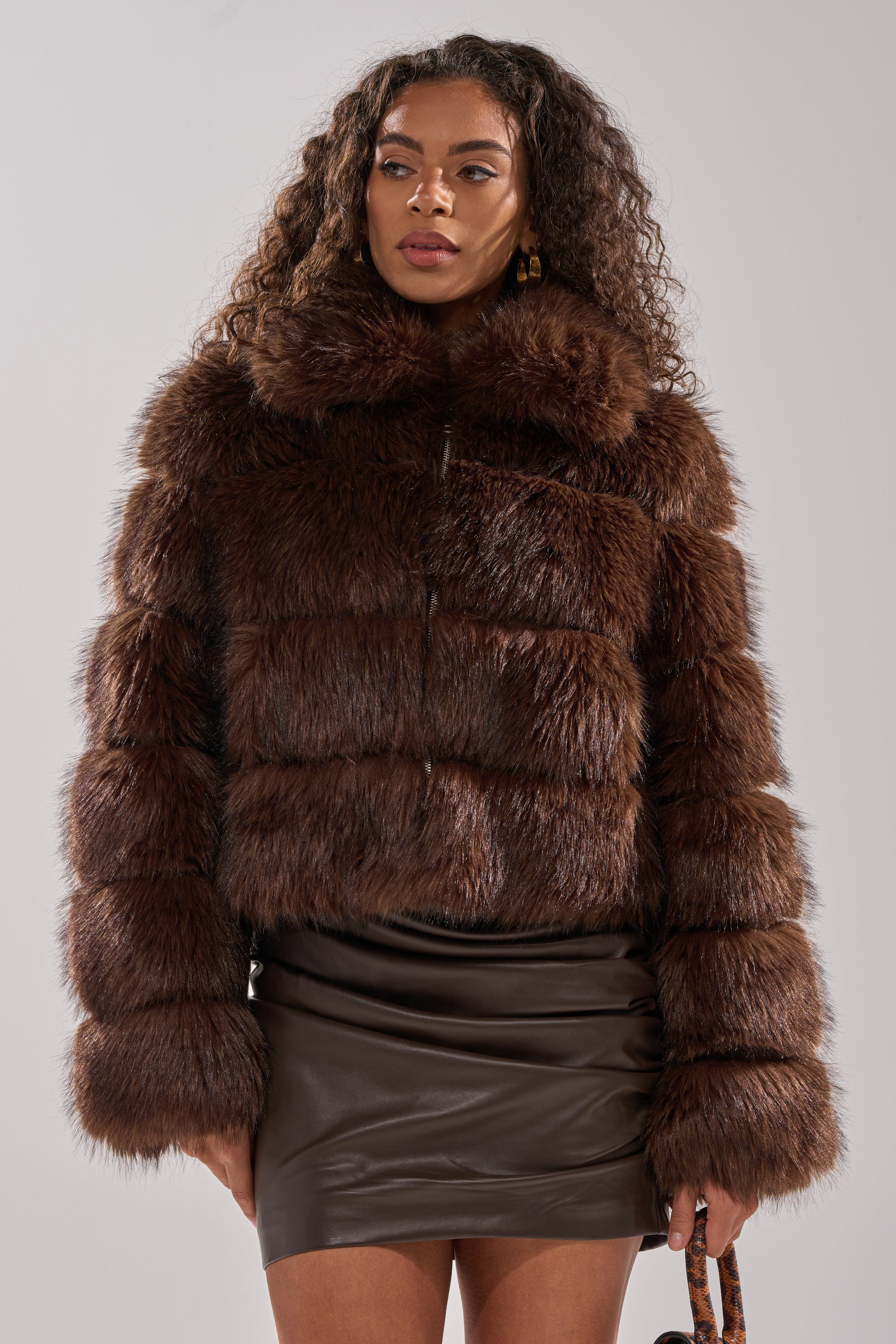 A woman with long curly hair wears the GOOD KARMA FAUX FUR jacket and a dark brown leather mini skirt, standing against a plain light gray background.