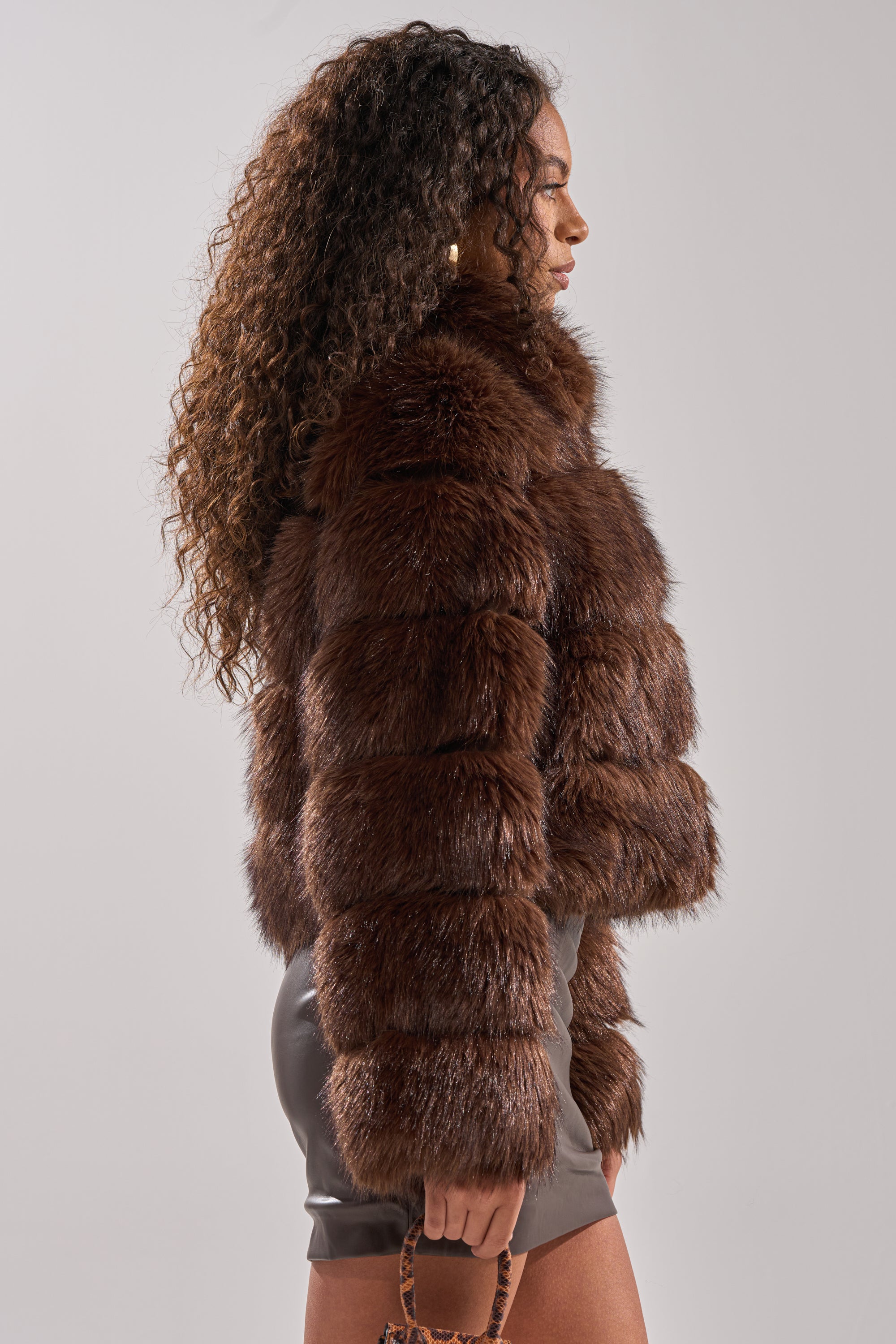 A woman with long, curly hair wears the GOOD KARMA FAUX FUR jacket featuring a thick, brown faux fur and oversized collar. She stands sideways against a plain, light background in a short skirt, holding a small bag.