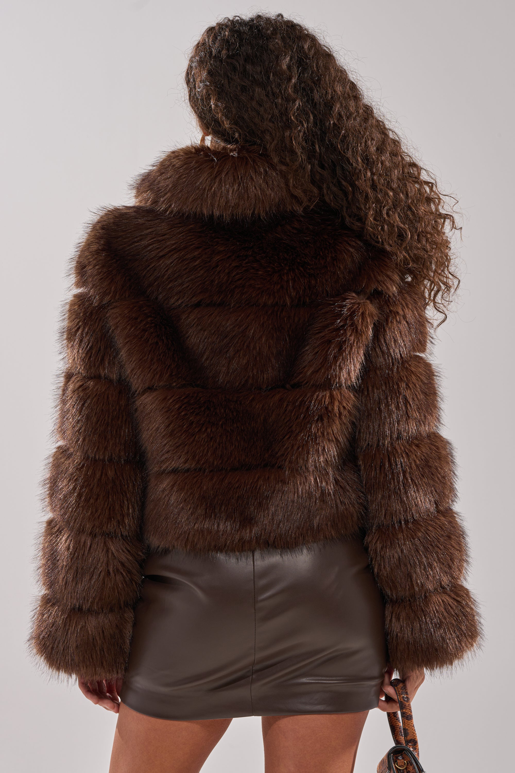 A woman with long curly hair, seen from behind, wears the GOOD KARMA FAUX FUR jacket with an oversized collar and a brown leather mini skirt. She holds a small bag in her left hand against a plain light background.