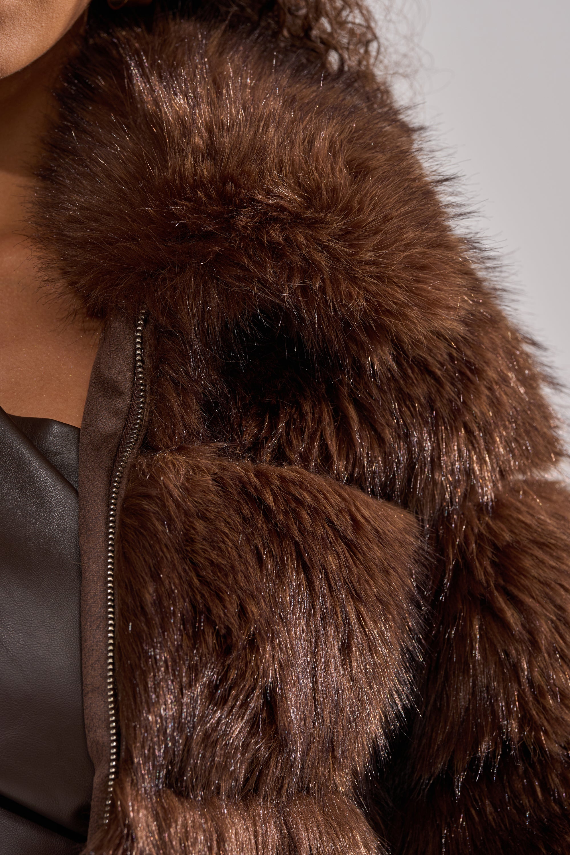 A person wears the GOOD KARMA FAUX FUR jacket in dark brown, featuring an oversized collar and visible zipper. The coat's soft, shiny faux fur stands out, with a hint of a dark leather top peeking underneath.