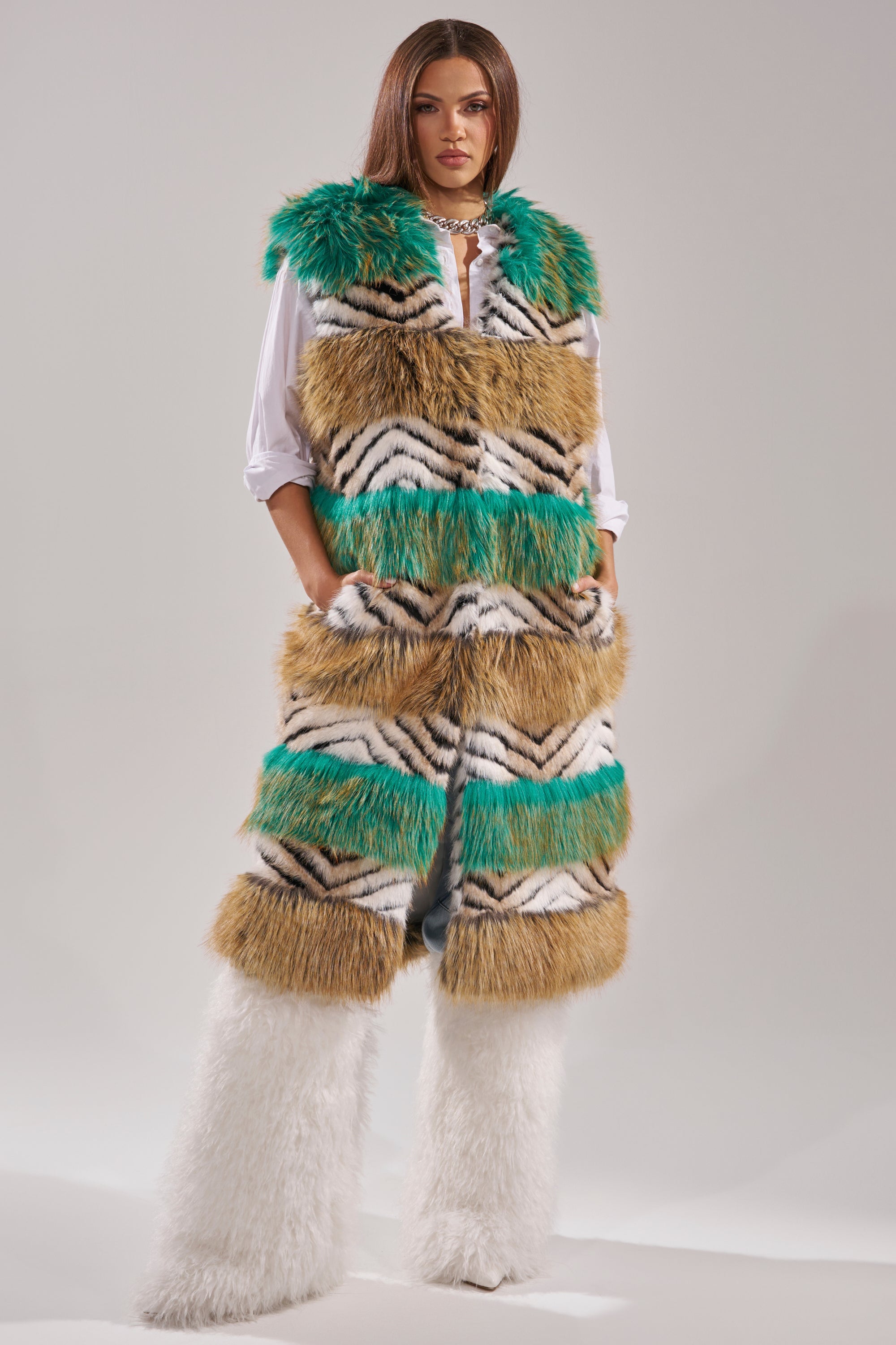 A woman exudes confidence in a winter luxe look, layering the HYDE PARK FAUX FUR VEST with bold green, tan, black, and white stripes over a white shirt and matching fluffy white pants against a simple backdrop. 0