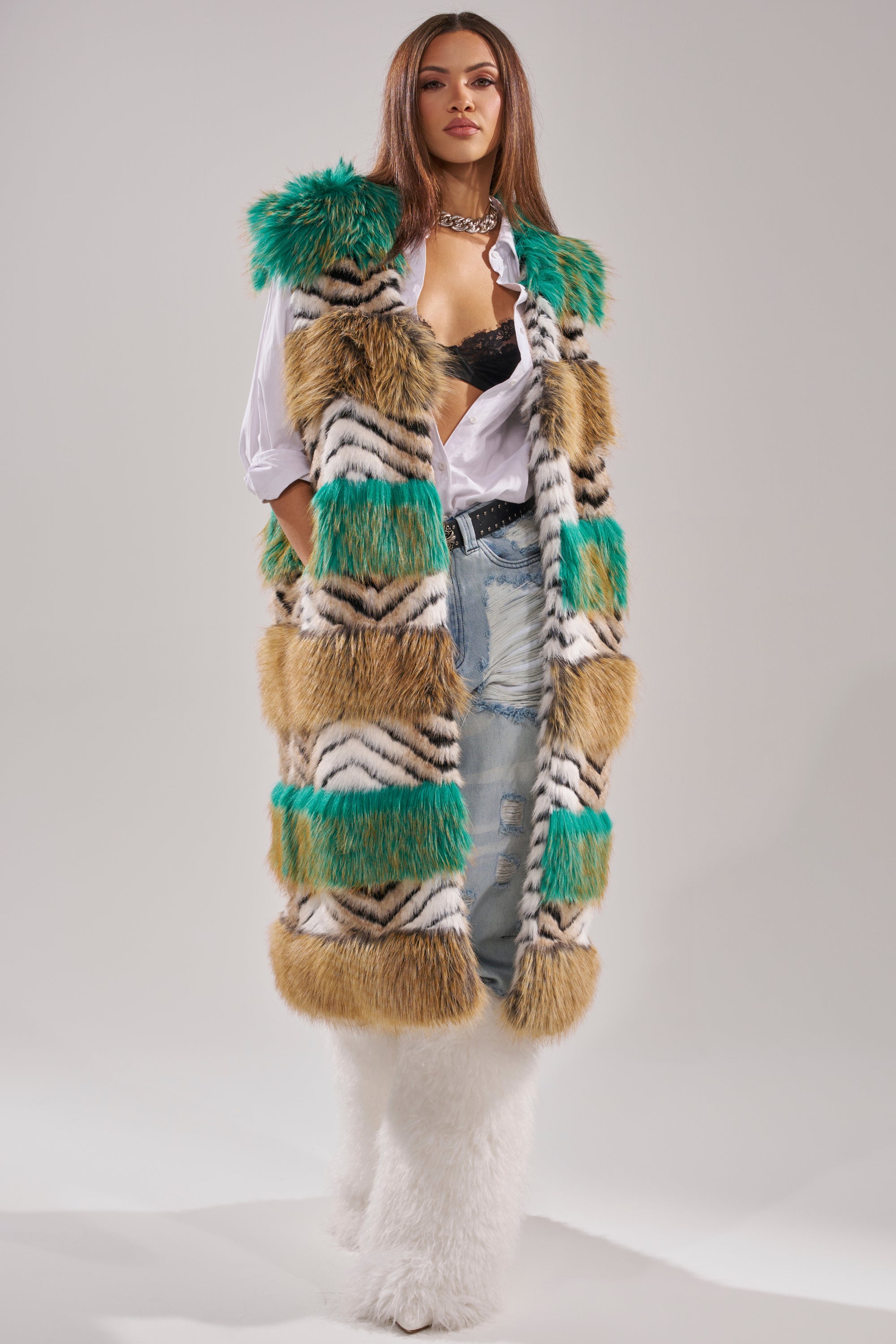 A woman exudes confidence in a winter luxury look, featuring the HYDE PARK FAUX FUR VEST with green, tan, and white stripes, styled over a white shirt and black bra, ripped jeans, chunky necklace, and fluffy white boots. 4