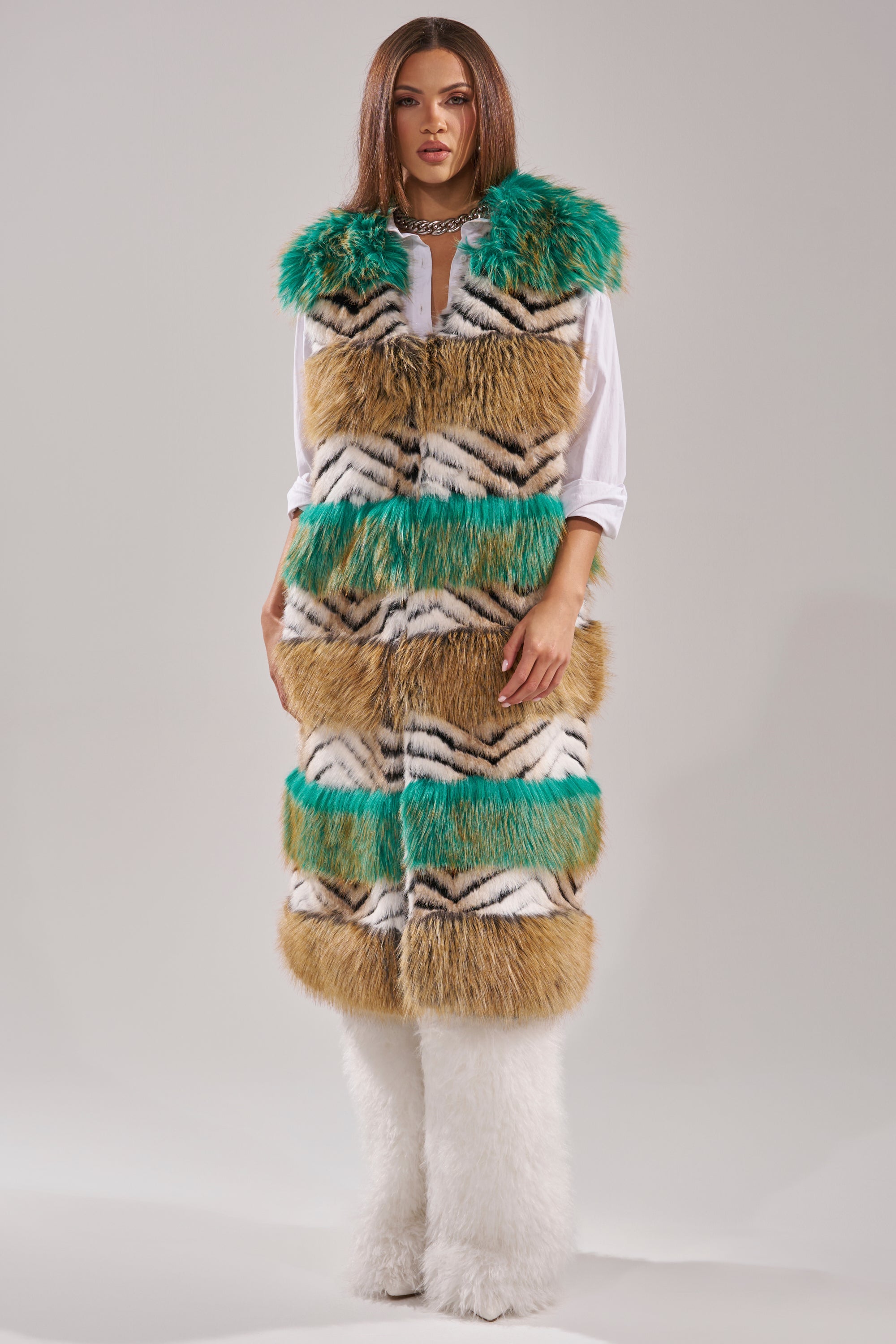 A woman models the HYDE PARK FAUX FUR VEST with brown, white, black, and green stripes over a white shirt, styled with white furry pants and a chunky chain necklace for an effortlessly luxurious winter look against a plain background. 5