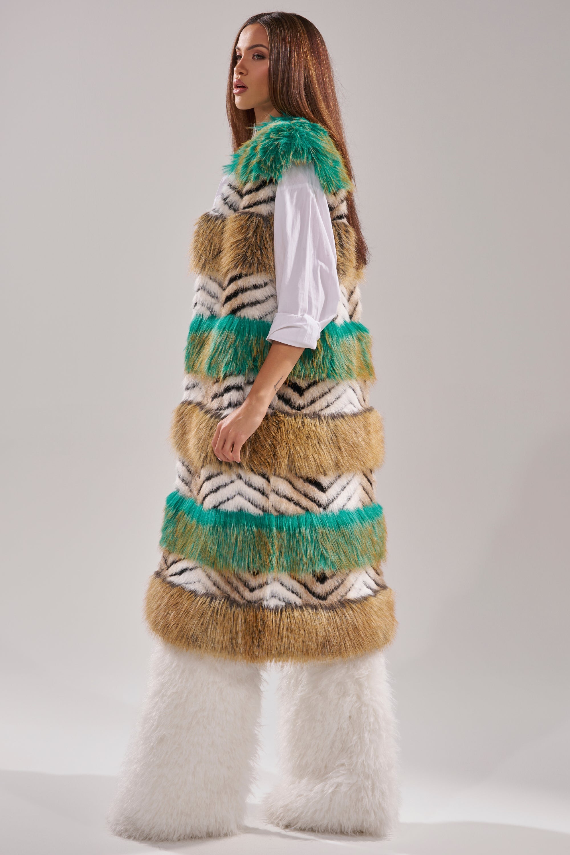 A woman poses in a studio wearing the HYDE PARK FAUX FUR VEST, which features green, tan, black, and white stripes over a white shirt and fluffy white wide-leg pants. 7