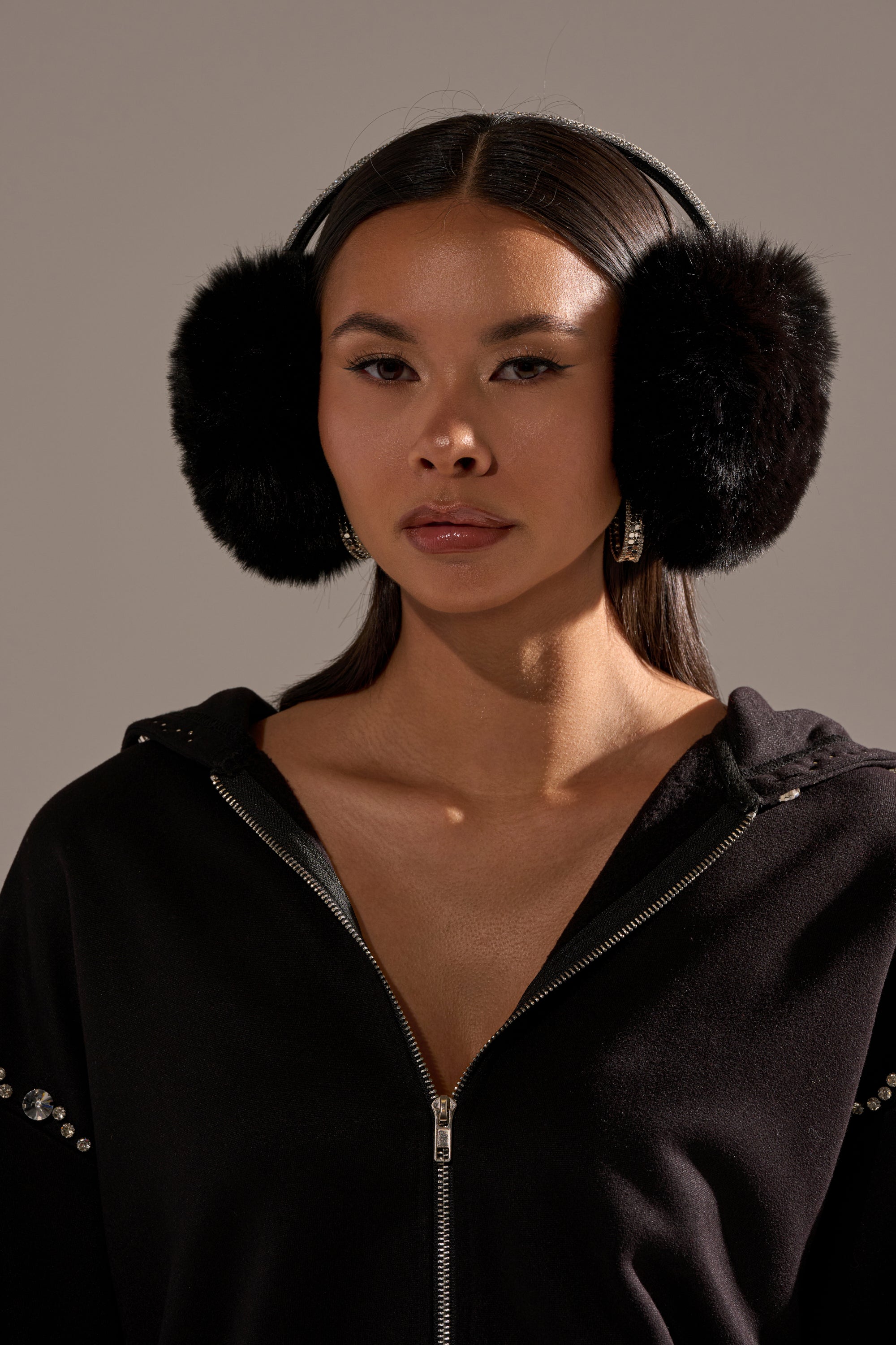 A woman with long dark hair poses against a plain background wearing EXTRA RHINESTONE EAR MUFFS IN BLACK and a black zip-up jacket with rhinestone details. 0