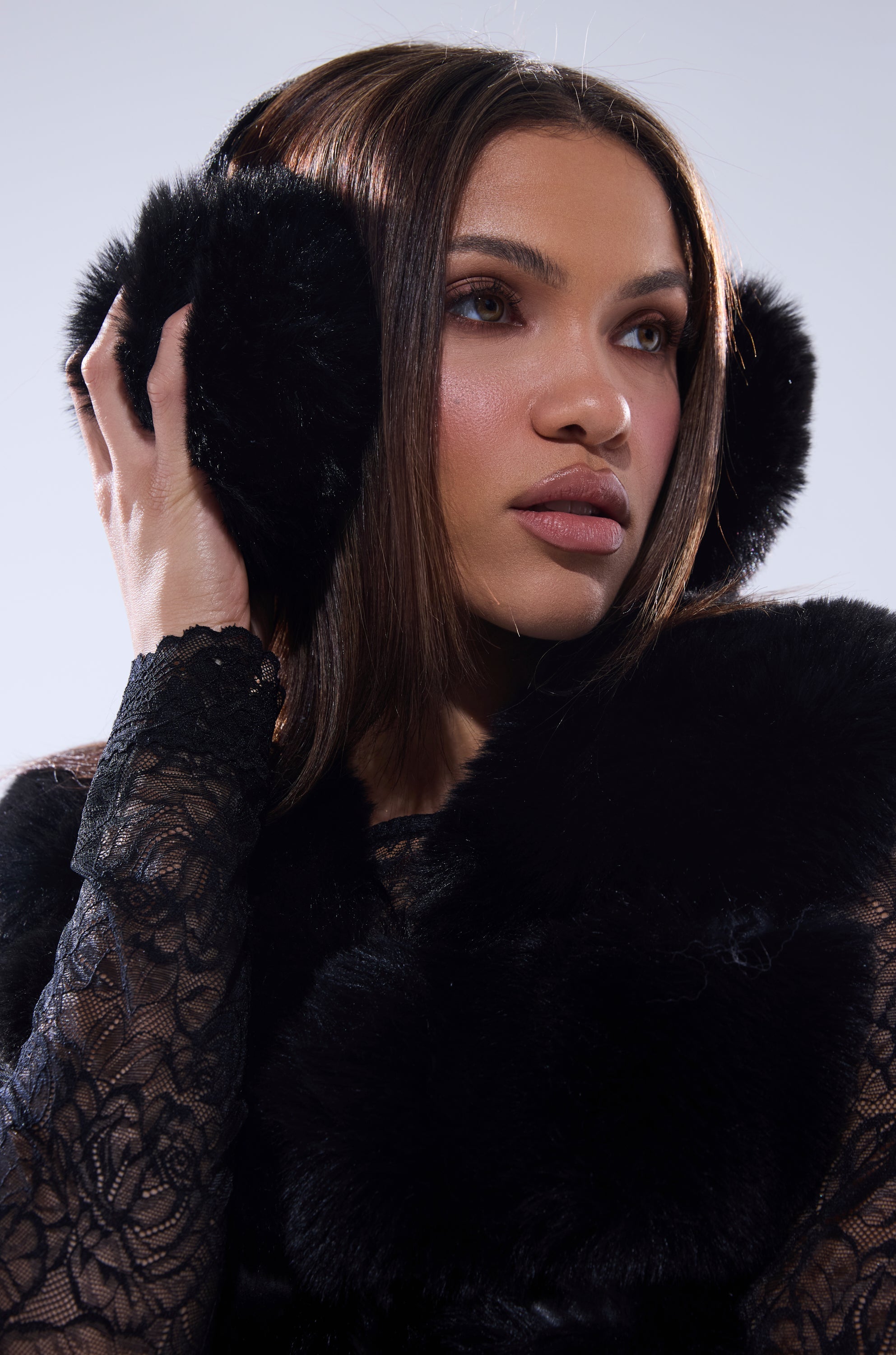A woman with brown, center-parted hair wears EXTRA RHINESTONE EAR MUFFS IN BLACK and a black lace top. She touches one earmuff while looking to the side against a softly lit, plain background, highlighting this chic winter accessory. 4