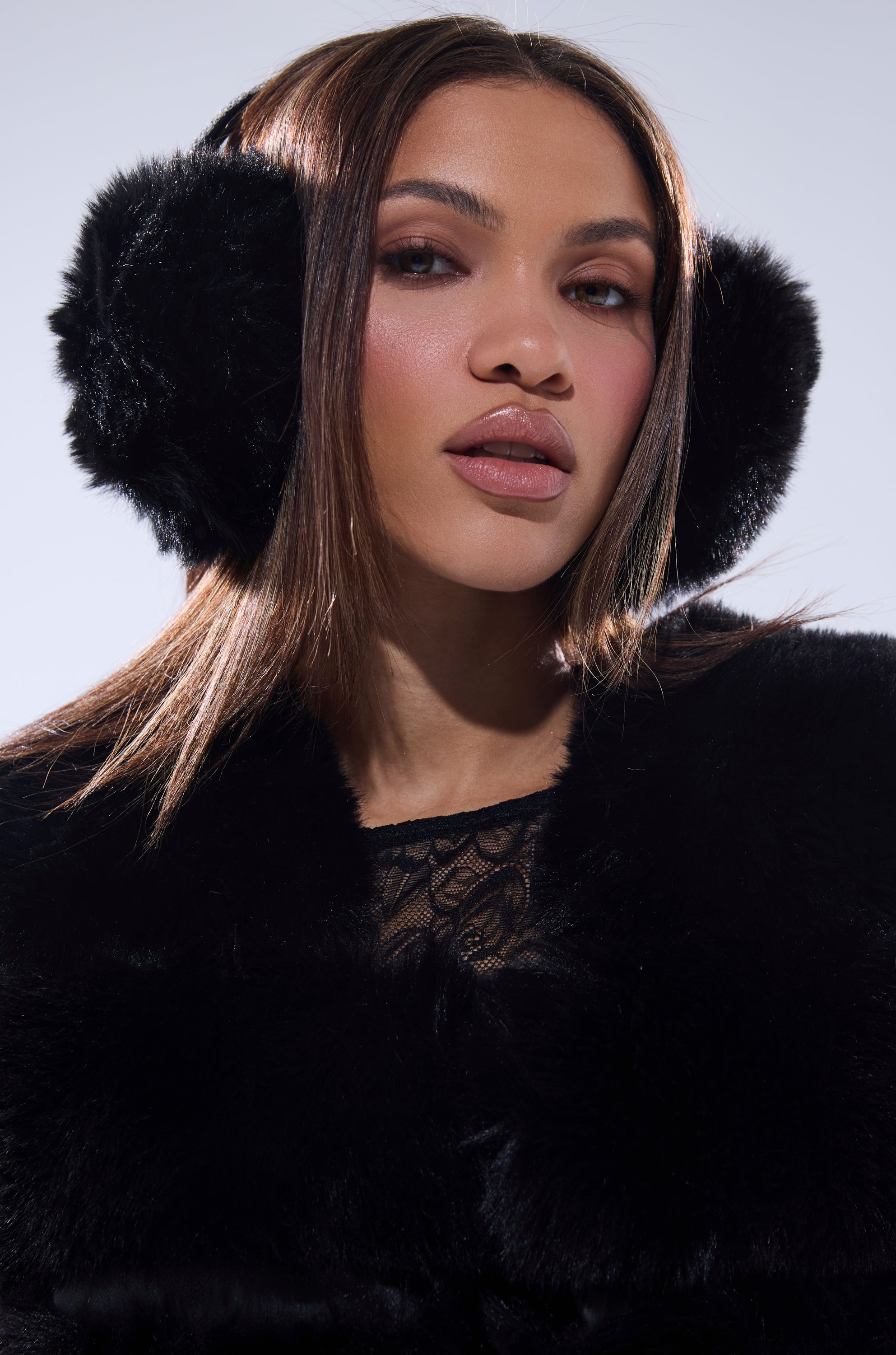 A woman with straight brown hair models the EXTRA RHINESTONE EAR MUFFS IN BLACK, styled with a black lace top and black fur coat against a light background—a chic winter look for accessory enthusiasts.