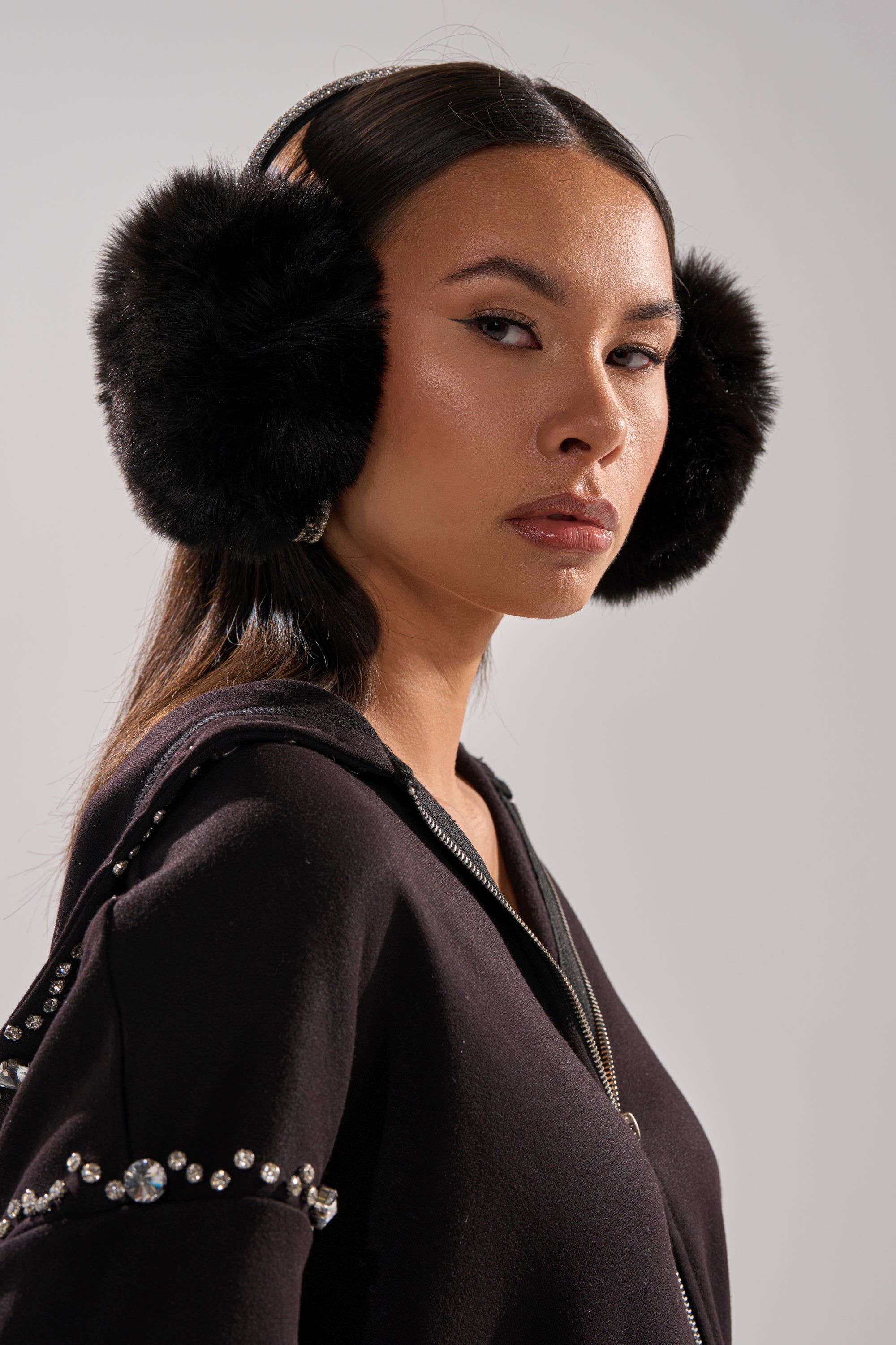 A woman with long dark hair wears EXTRA RHINESTONE EAR MUFFS IN BLACK and a black rhinestone hoodie, confidently posing against a plain background—making a bold, stylish winter statement.