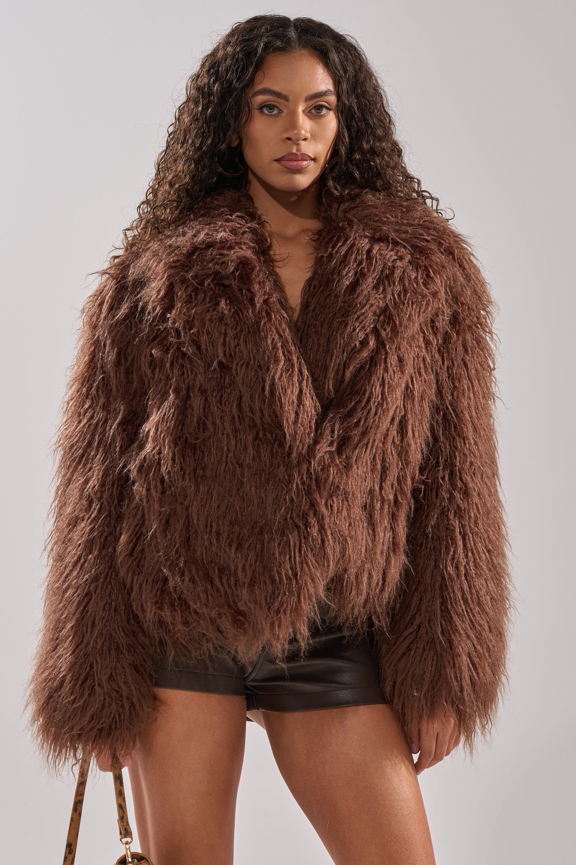 A woman with curly hair models the SHORT N SWEET FAUX FUR COAT in chocolate, paired with black leather shorts and a small handbag, standing against a plain light background.