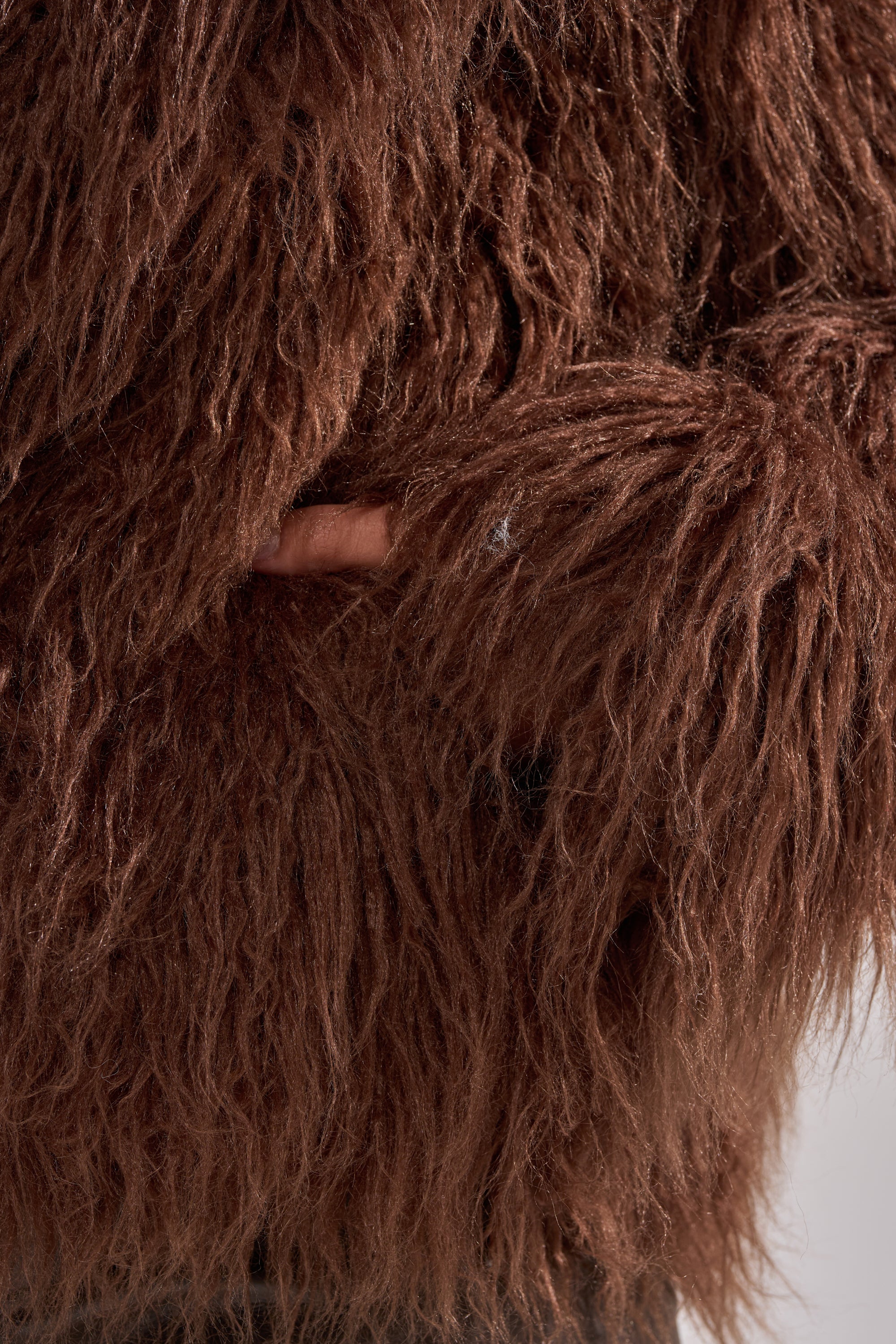 A close-up of the SHORT N SWEET FAUX FUR COAT in chocolate brown, featuring its shaggy faux fur texture and a person's hand partially tucked into the side pocket.