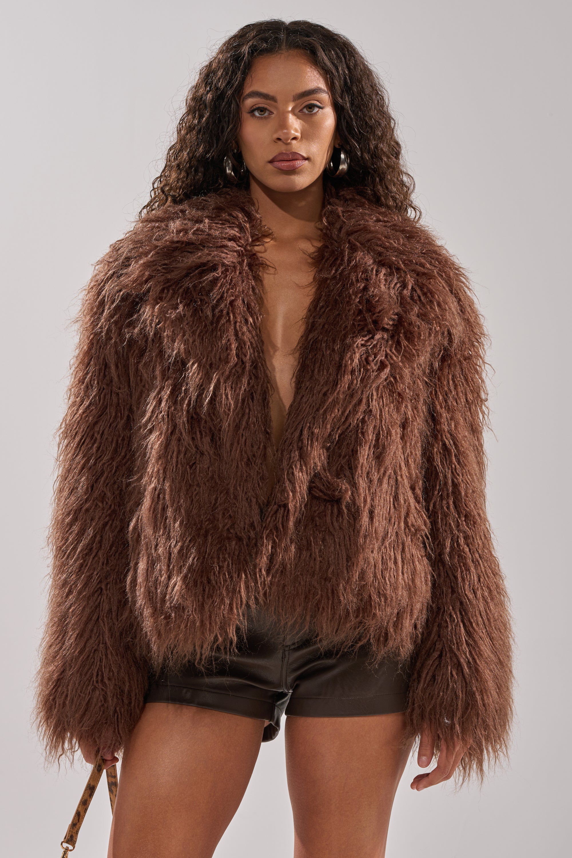 A woman with long, curly hair models the SHORT N SWEET FAUX FUR COAT in chocolate over black leather shorts, standing against a plain light gray background.