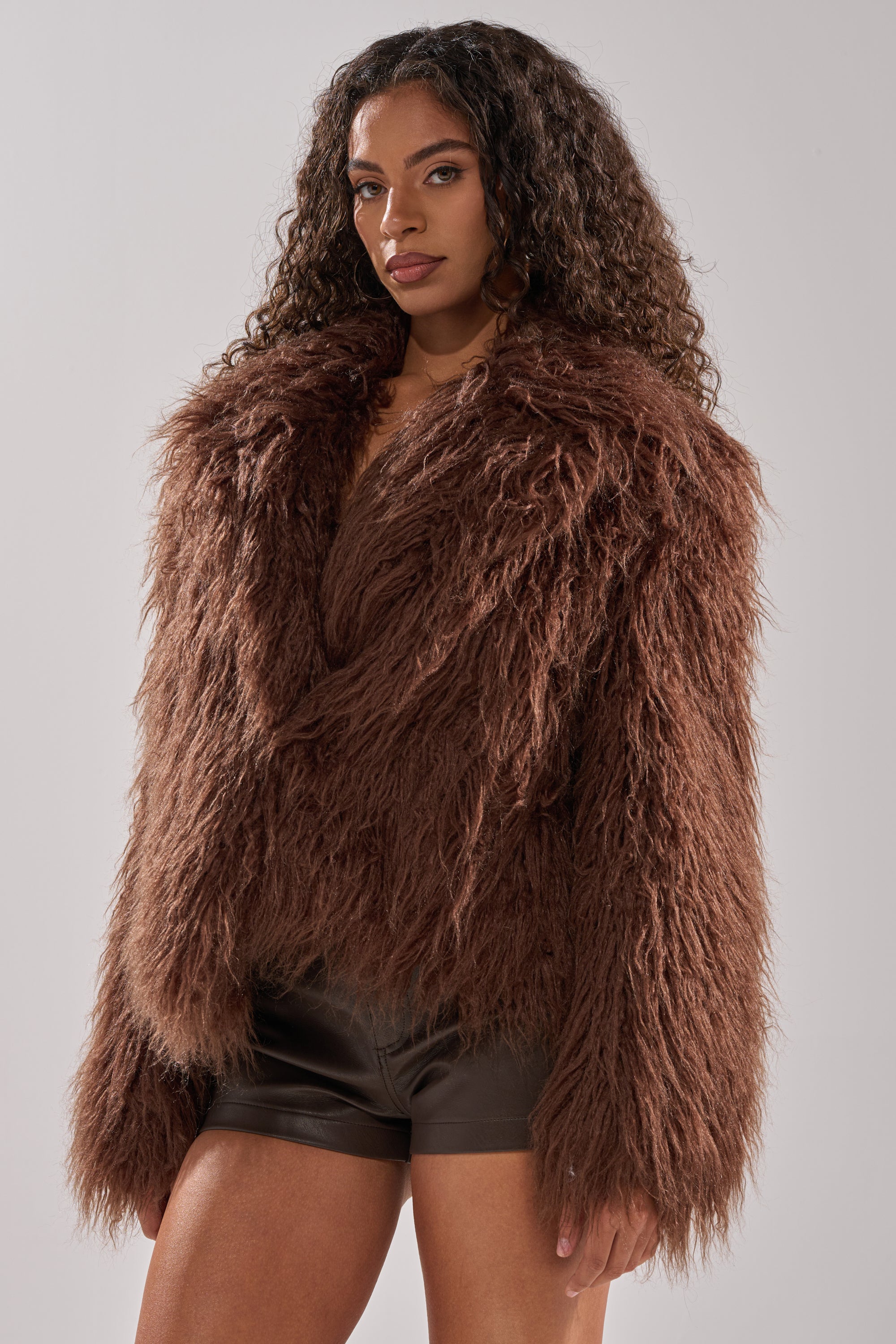 A woman with long curly hair wears the SHORT N SWEET FAUX FUR COAT in chocolate over dark brown shorts, standing against a plain light gray background.