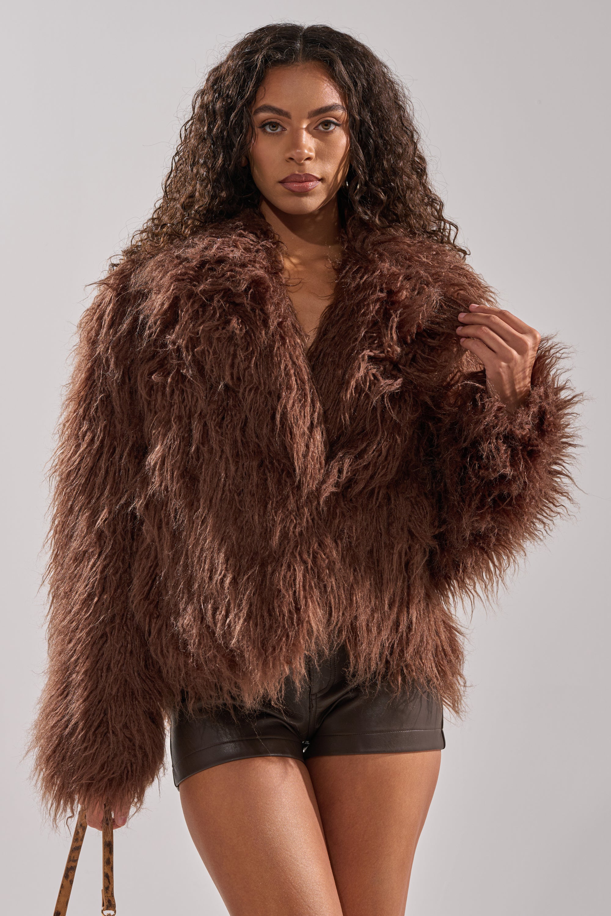 A woman with long curly hair models the SHORT N SWEET FAUX FUR COAT in chocolate, paired with black shorts and a bag, standing against a plain light gray background.
