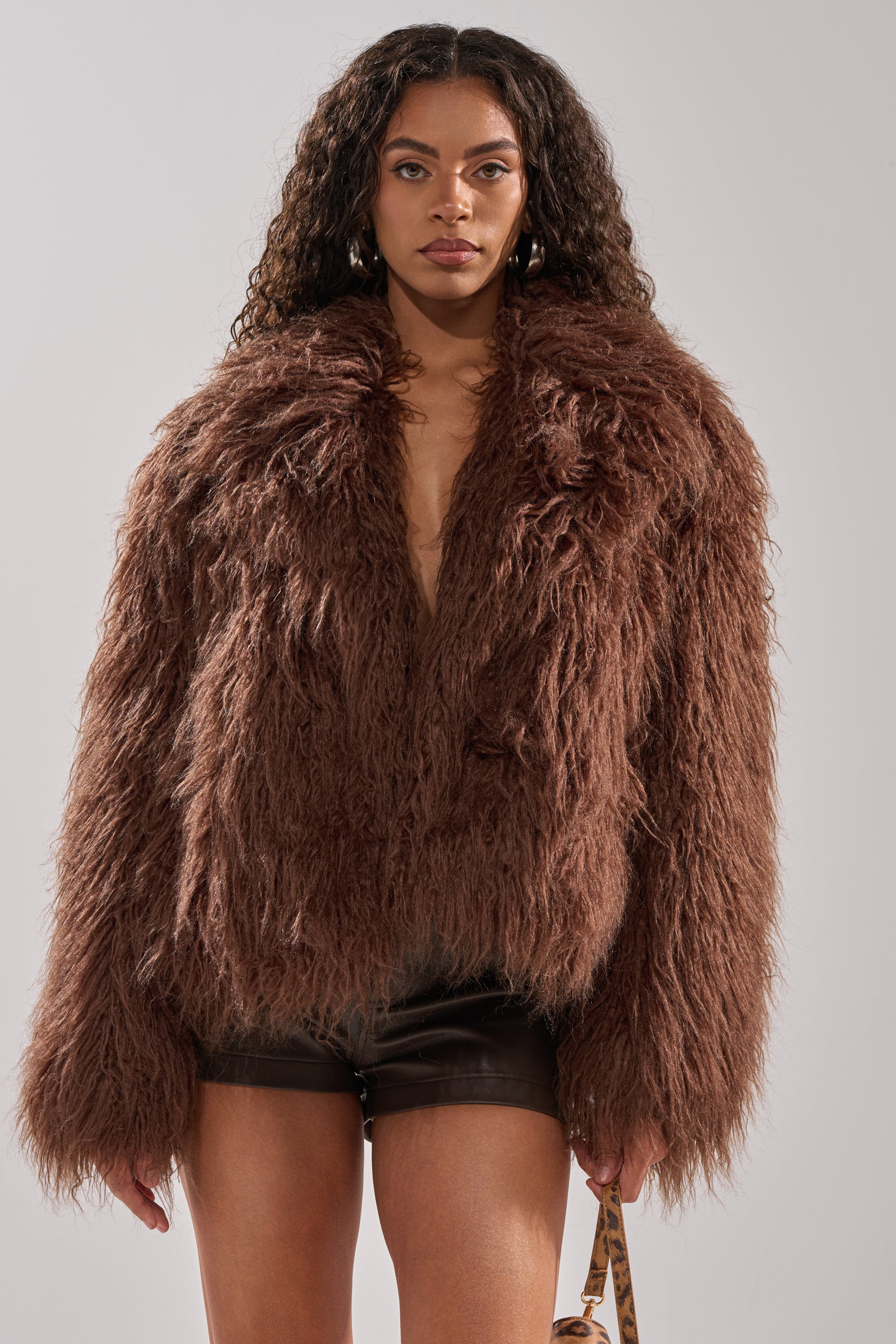 A woman with long curly hair wears the SHORT N SWEET FAUX FUR COAT in chocolate over black shorts, standing against a plain light gray background.
