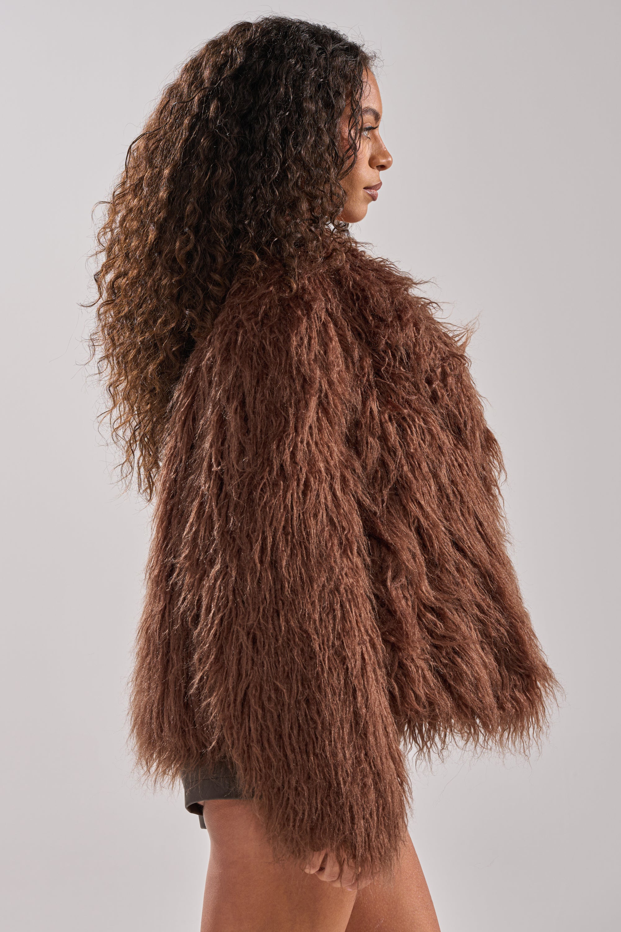 A woman with long, curly hair stands in profile wearing the SHORT N SWEET FAUX FUR COAT in chocolate brown, set against a plain light gray background.