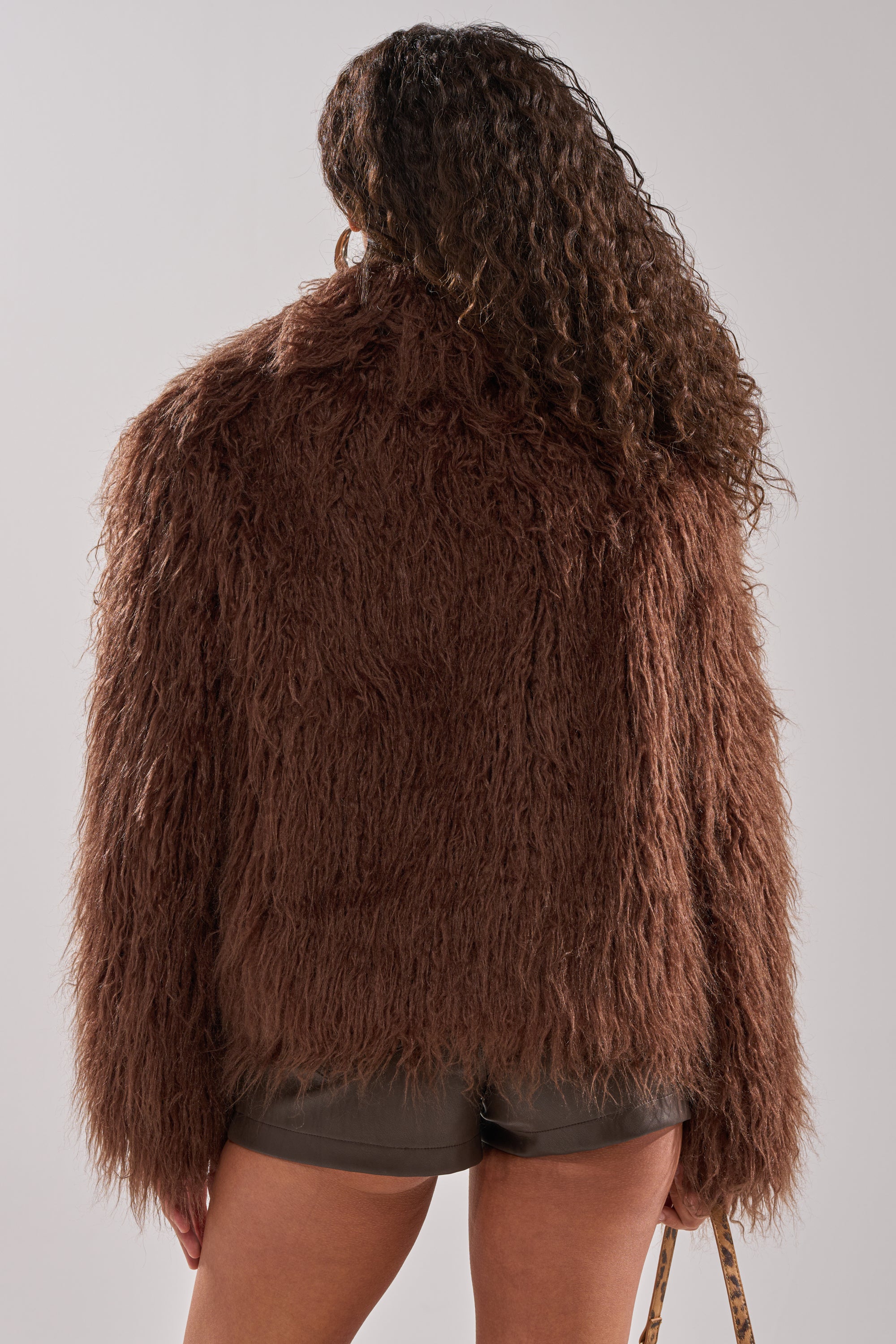 A person with long, curly hair is shown from the back in a chocolate SHORT N SWEET FAUX FUR COAT and dark shorts, holding a bag in their left hand against a plain background.
