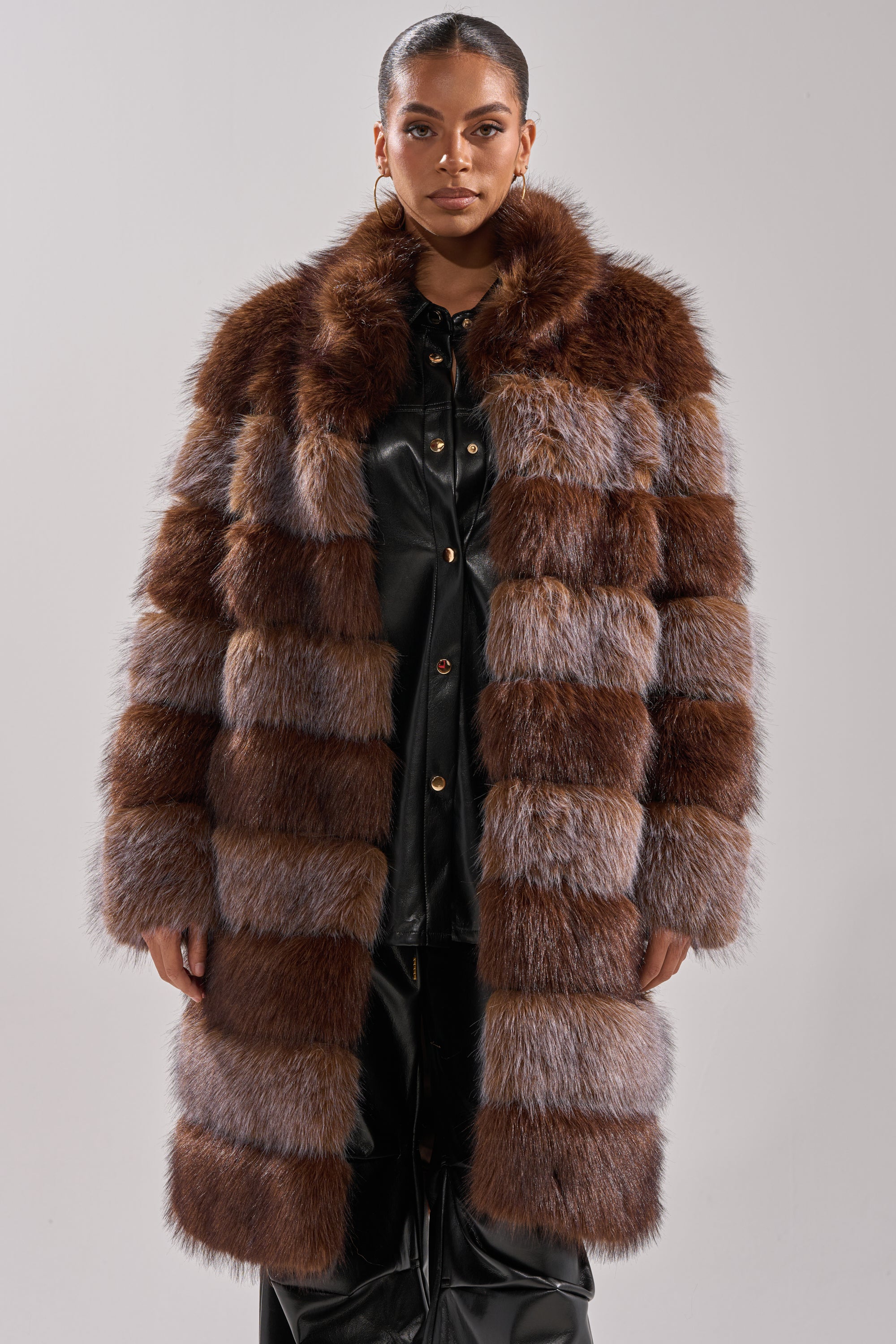 A woman faces forward in the LITTLE BIT OF SPICE PANELED FAUX FUR COAT, a luxurious brown-and-gray longline faux fur piece styled over a black leather outfit. She wears hoop earrings with her hair pulled back, against a plain light gray background.