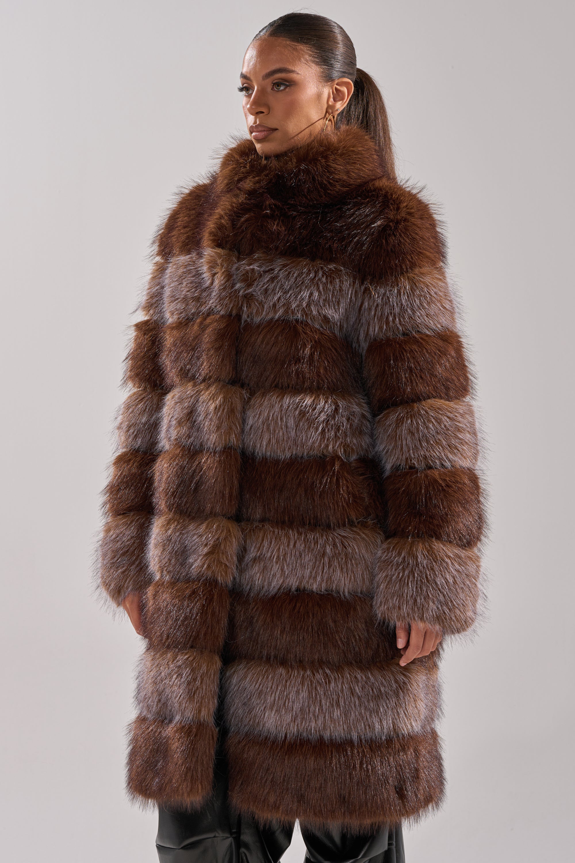 A woman confidently poses against a light-gray background, wearing the LITTLE BIT OF SPICE PANELED FAUX FUR COAT—a luxurious, knee-length striped brown and gray coat with a longline silhouette.