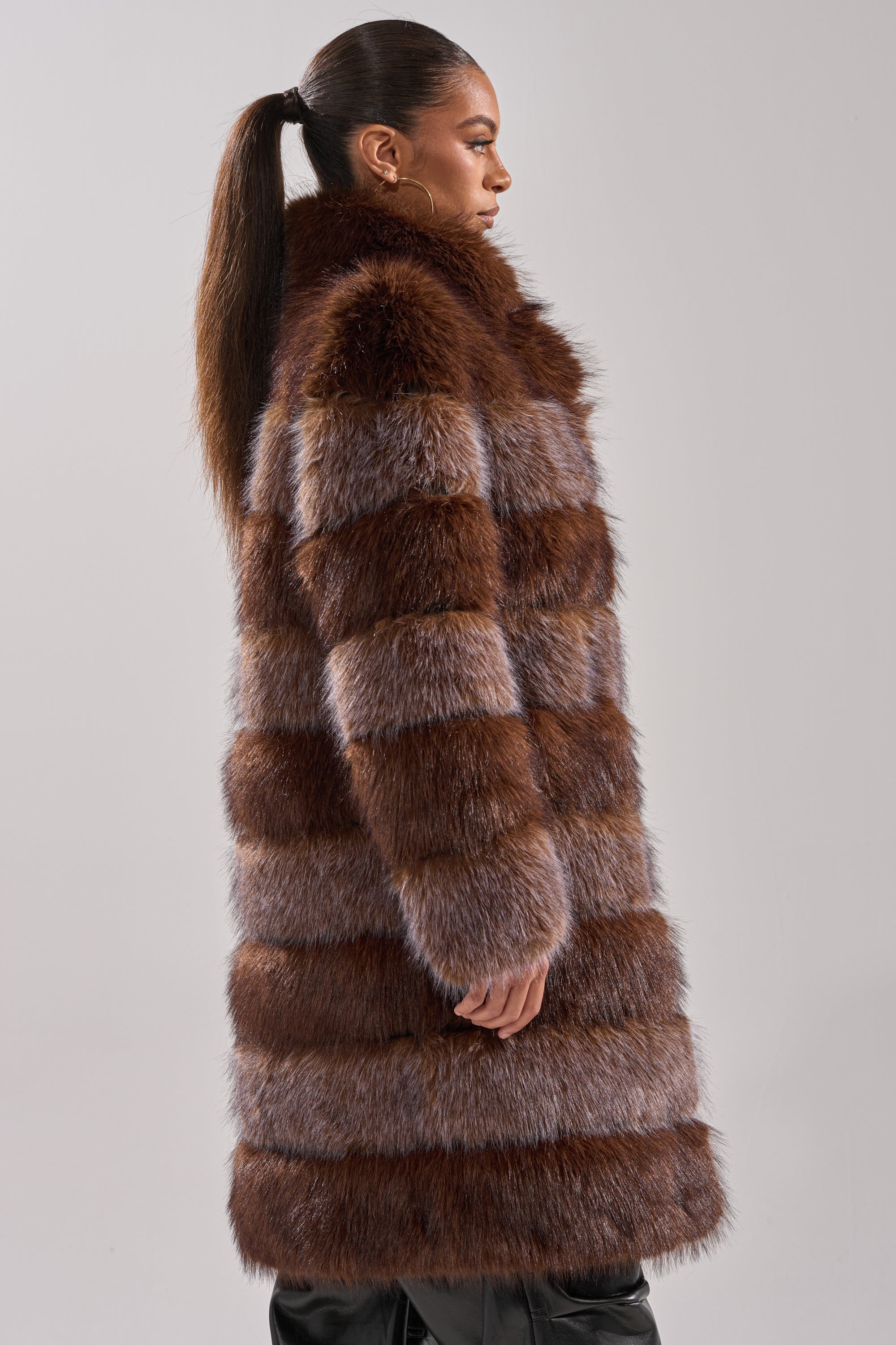 A woman with a high ponytail stands in profile against a plain light gray background, modeling the LITTLE BIT OF SPICE PANELED FAUX FUR COAT featuring brown and gray stripes.