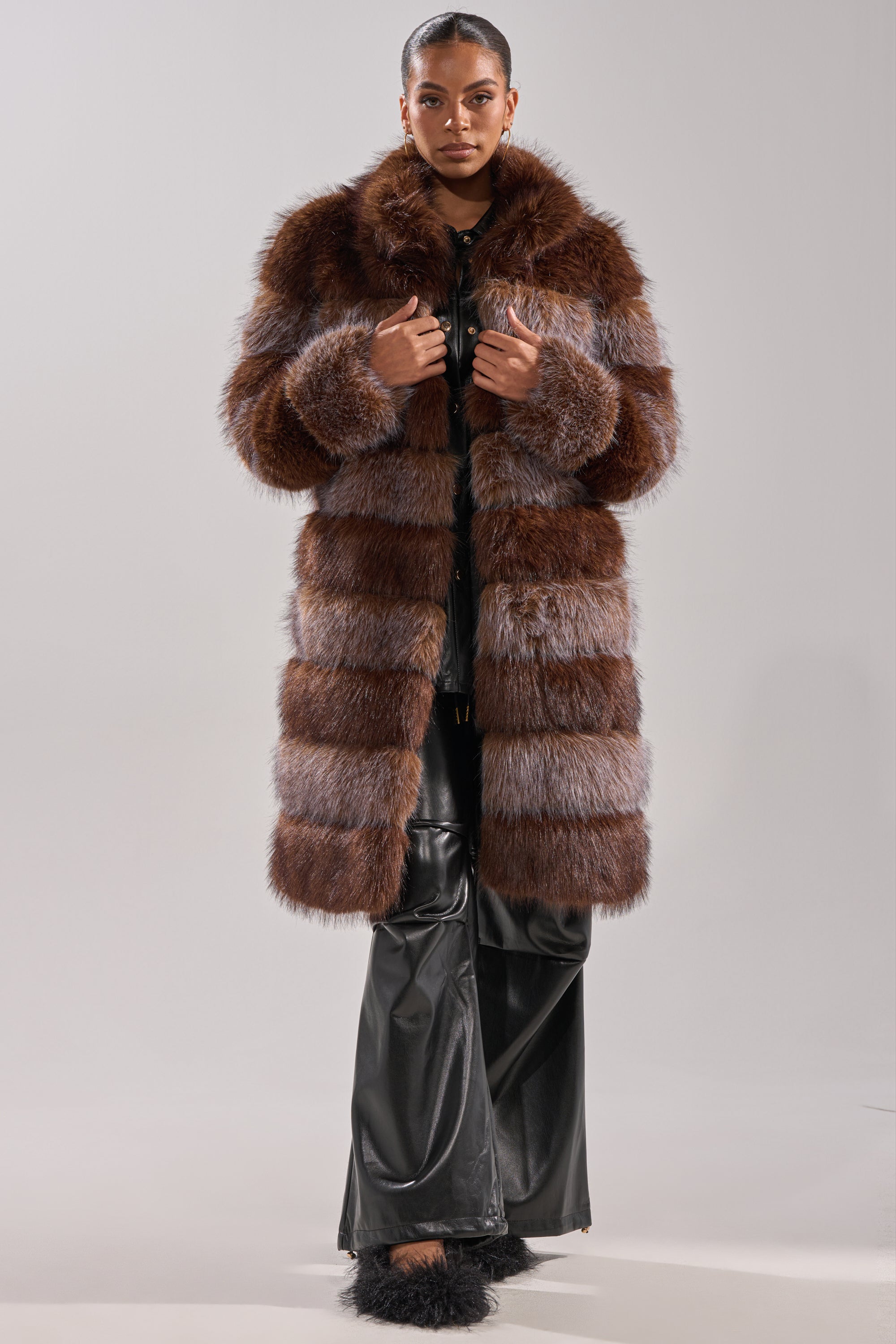 A person faces forward in the LITTLE BIT OF SPICE PANELED FAUX FUR COAT, featuring brown and gray stripes, styled with black leather pants and black furry shoes against a plain light gray background.