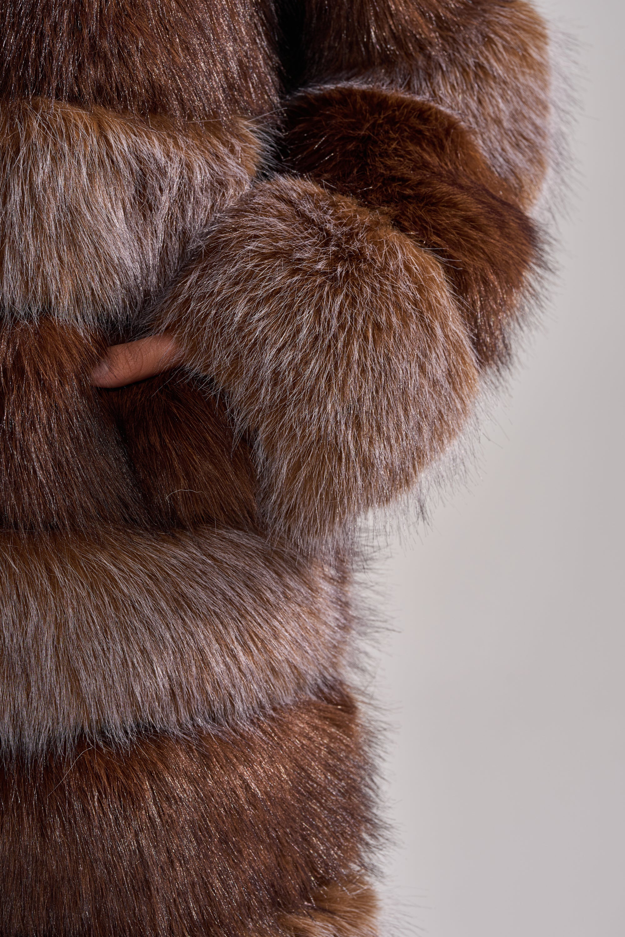 A person models the LITTLE BIT OF SPICE PANELED FAUX FUR COAT, a thick brown and gray longline piece with a soft, dense texture and paneled design, standing with one hand in the pocket against a neutral background.