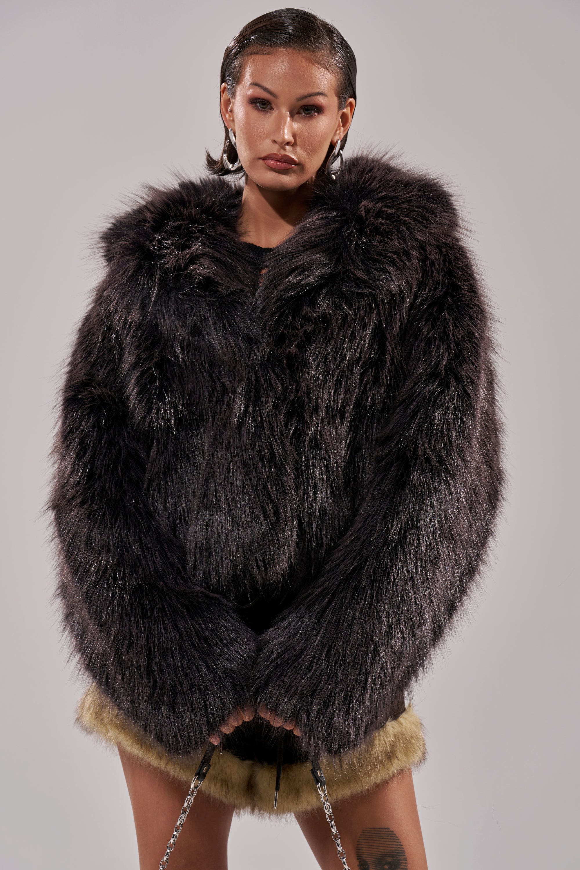 A person with slicked-back hair models the oversized DUSK TIL DAWN FAUX FUR COAT, featuring wide sleeves and tan furry trim, posing against a plain gray background. 0