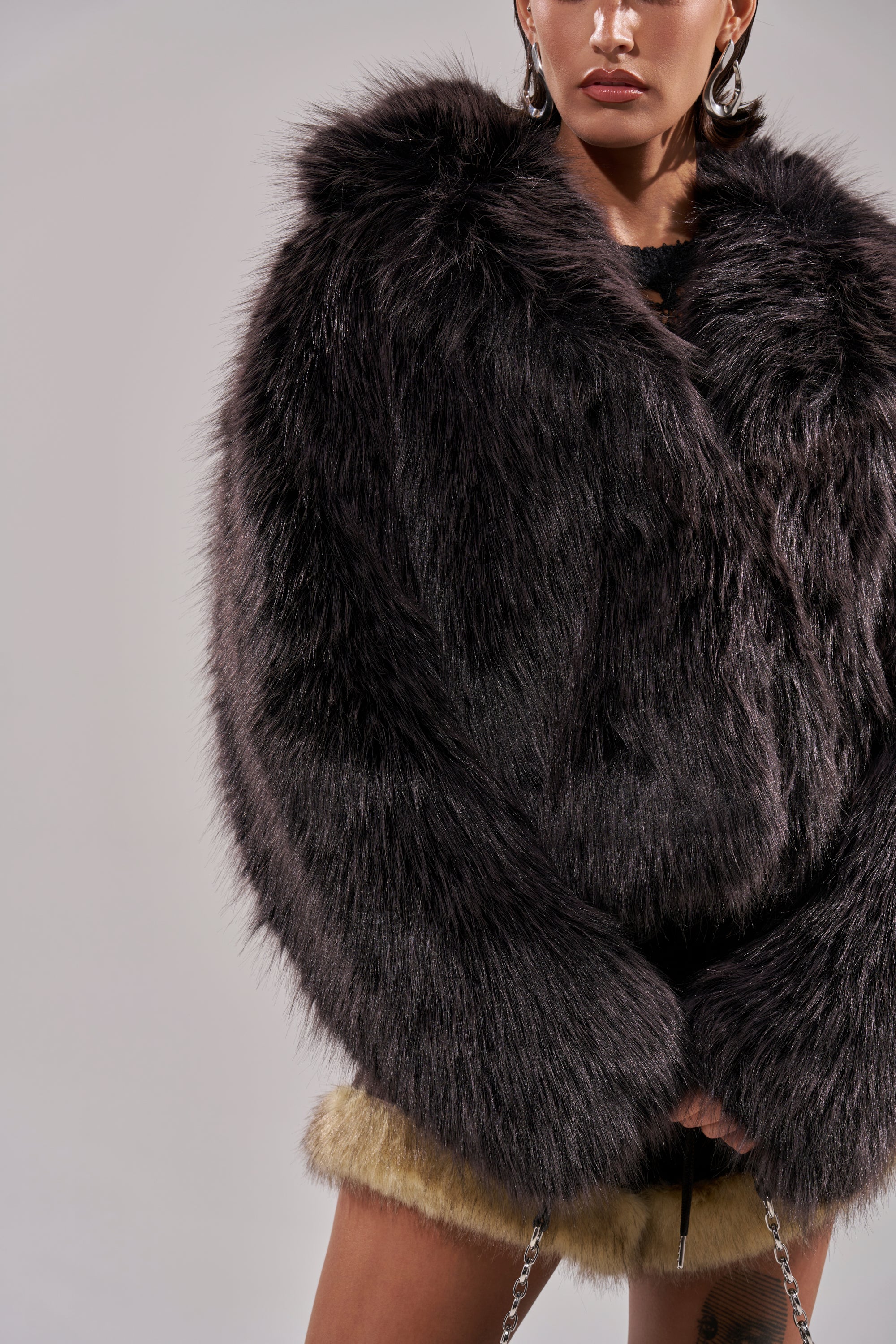 Wearing the AKIRA Label DUSK TIL DAWN FAUX FUR COAT and light furry shorts, a person holds a chain-strap accessory against a plain background, their face partially visible. 11