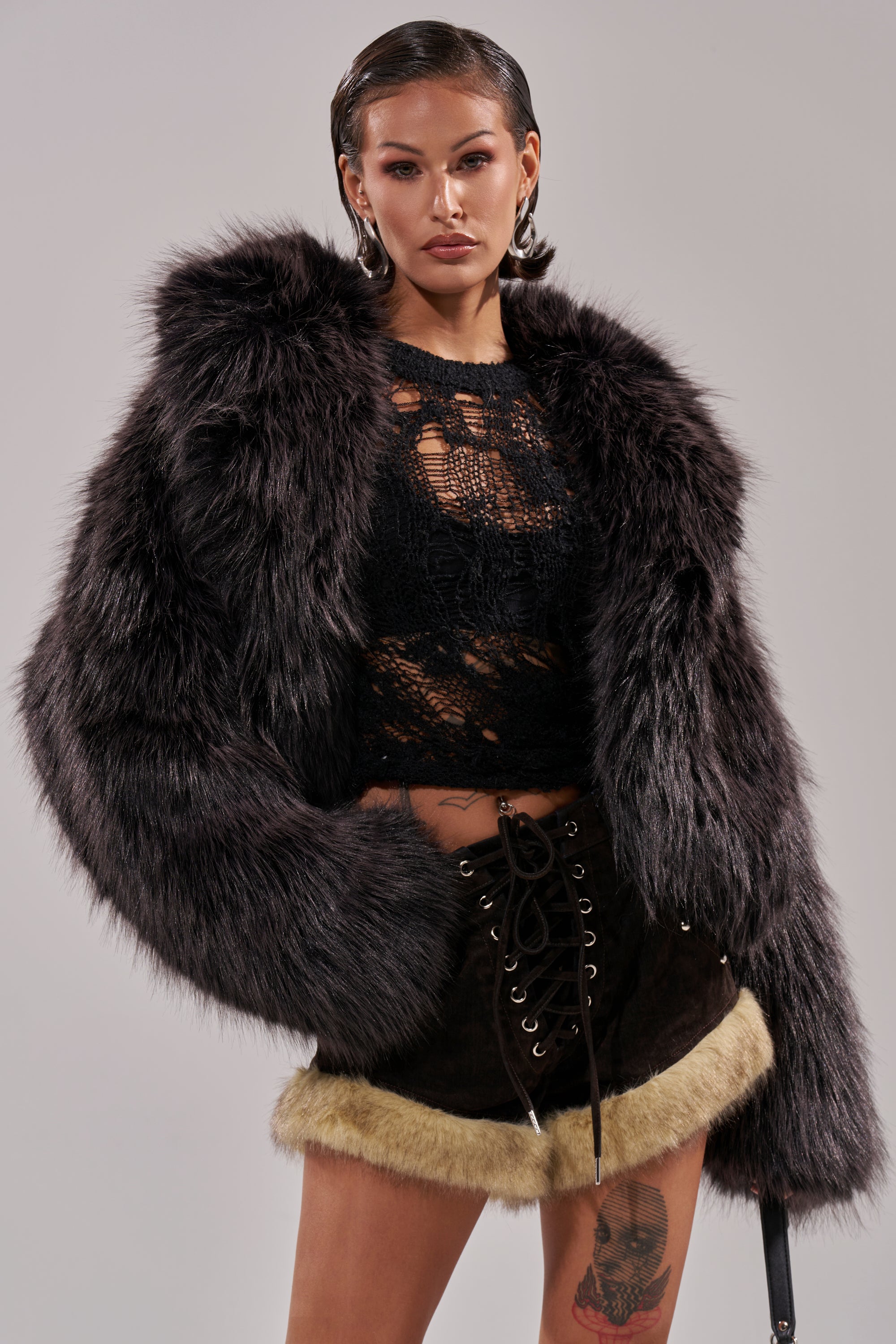 A woman with slicked-back hair confidently poses in the DUSK TIL DAWN FAUX FUR COAT, a black lace top, and black lace-up shorts with tan fur trim. She reveals a thigh tattoo against a plain background. 1