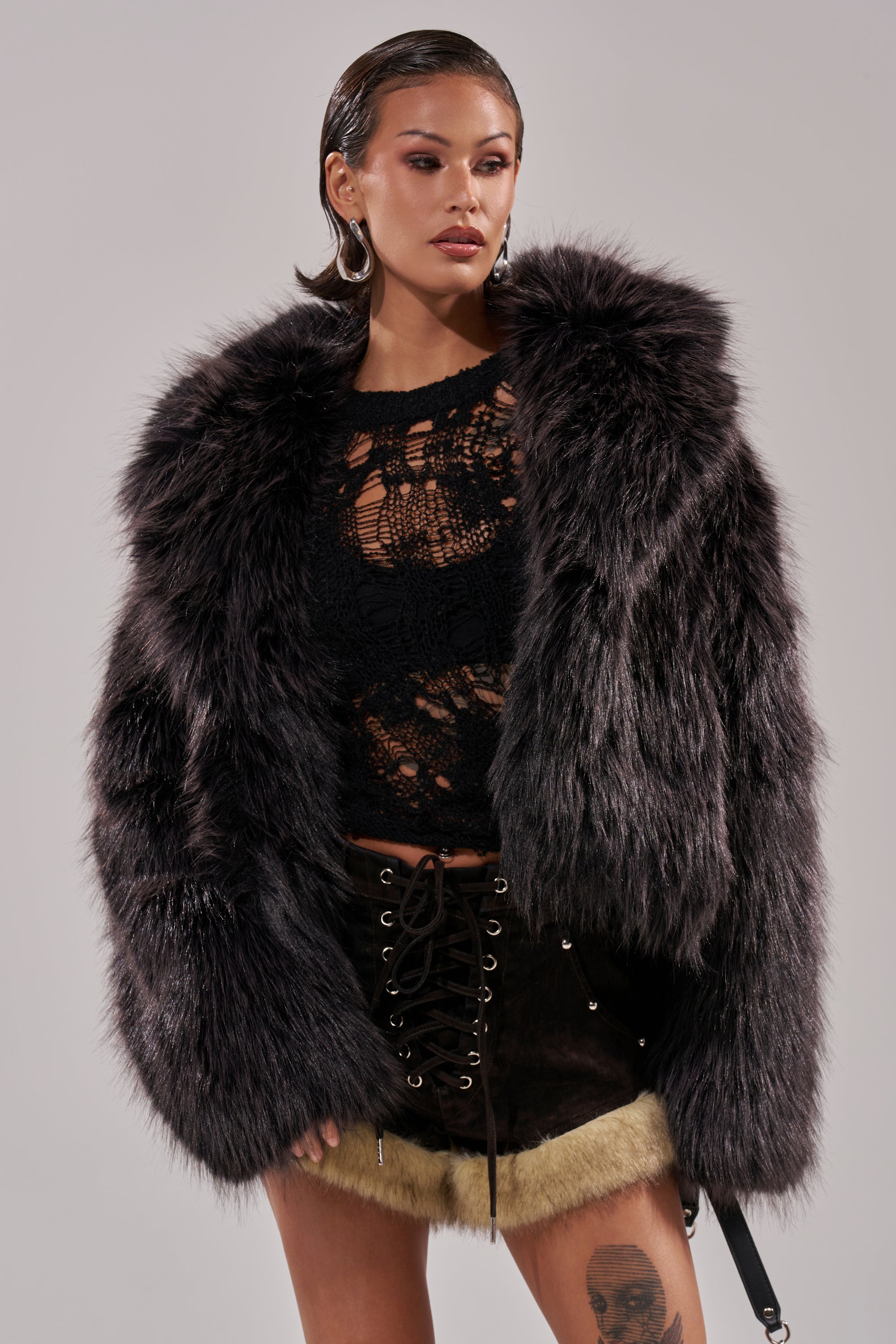 A woman wearing the DUSK TIL DAWN FAUX FUR COAT over a black lace top and lace-up mini skirt with beige fur trim poses against a plain background, her hair slicked back and makeup bold. 3